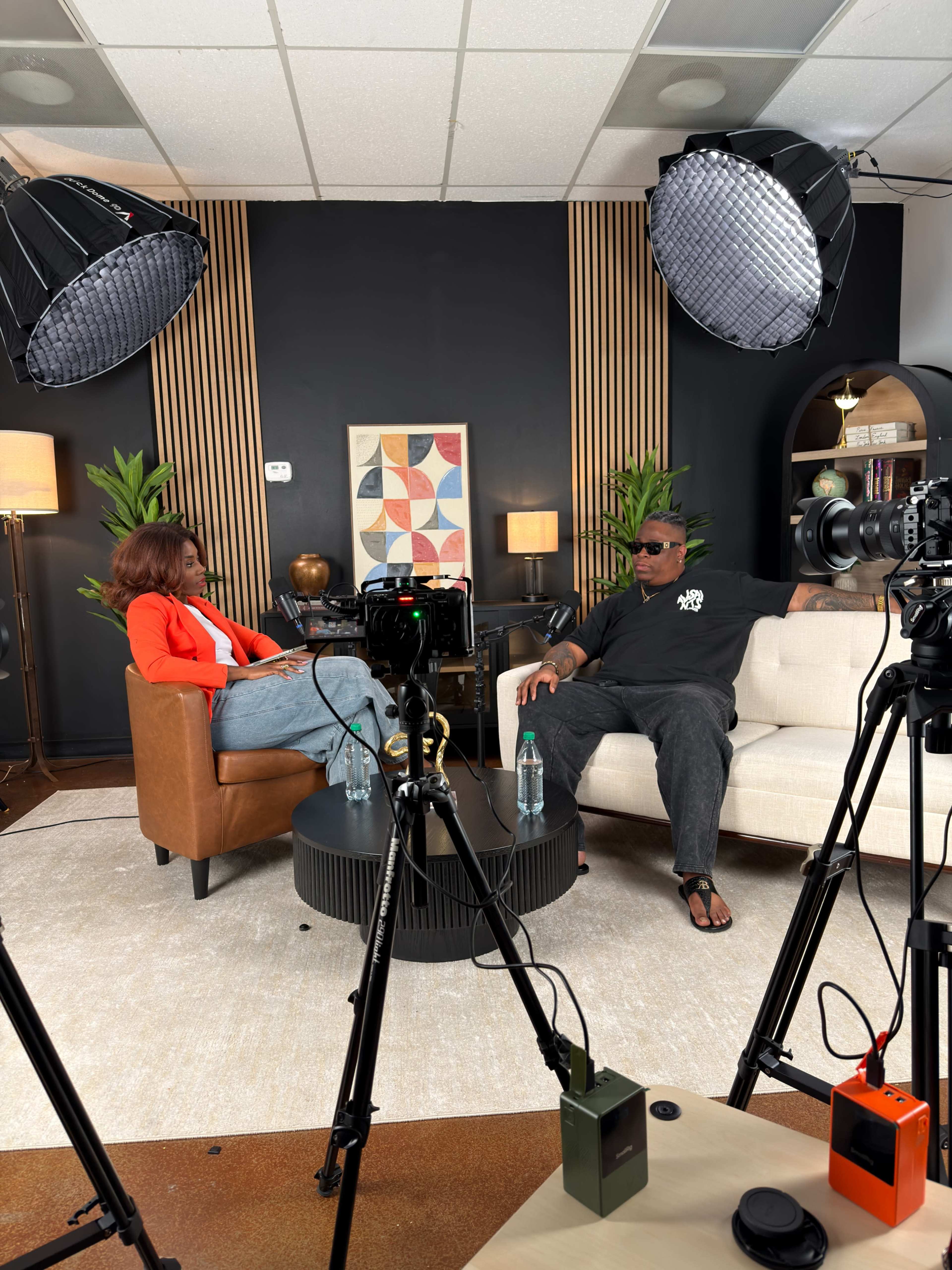 A man and a woman sit in a well-decorated studio setting, with cameras and lighting equipment positioned around them.