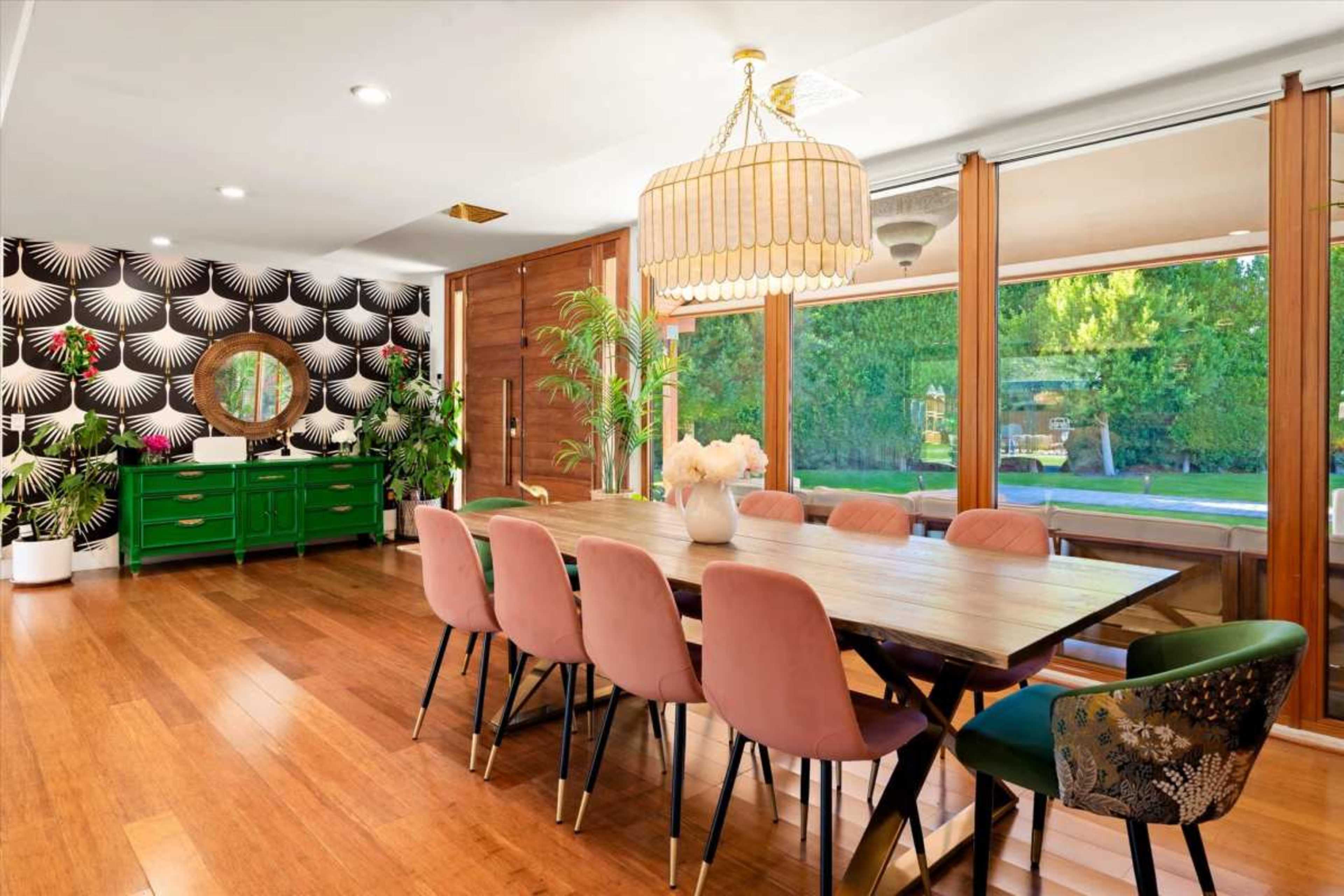 A modern dining room features a large wooden table surrounded by colorful chairs, with a decorative wallpaper and large windows providing natural light.
