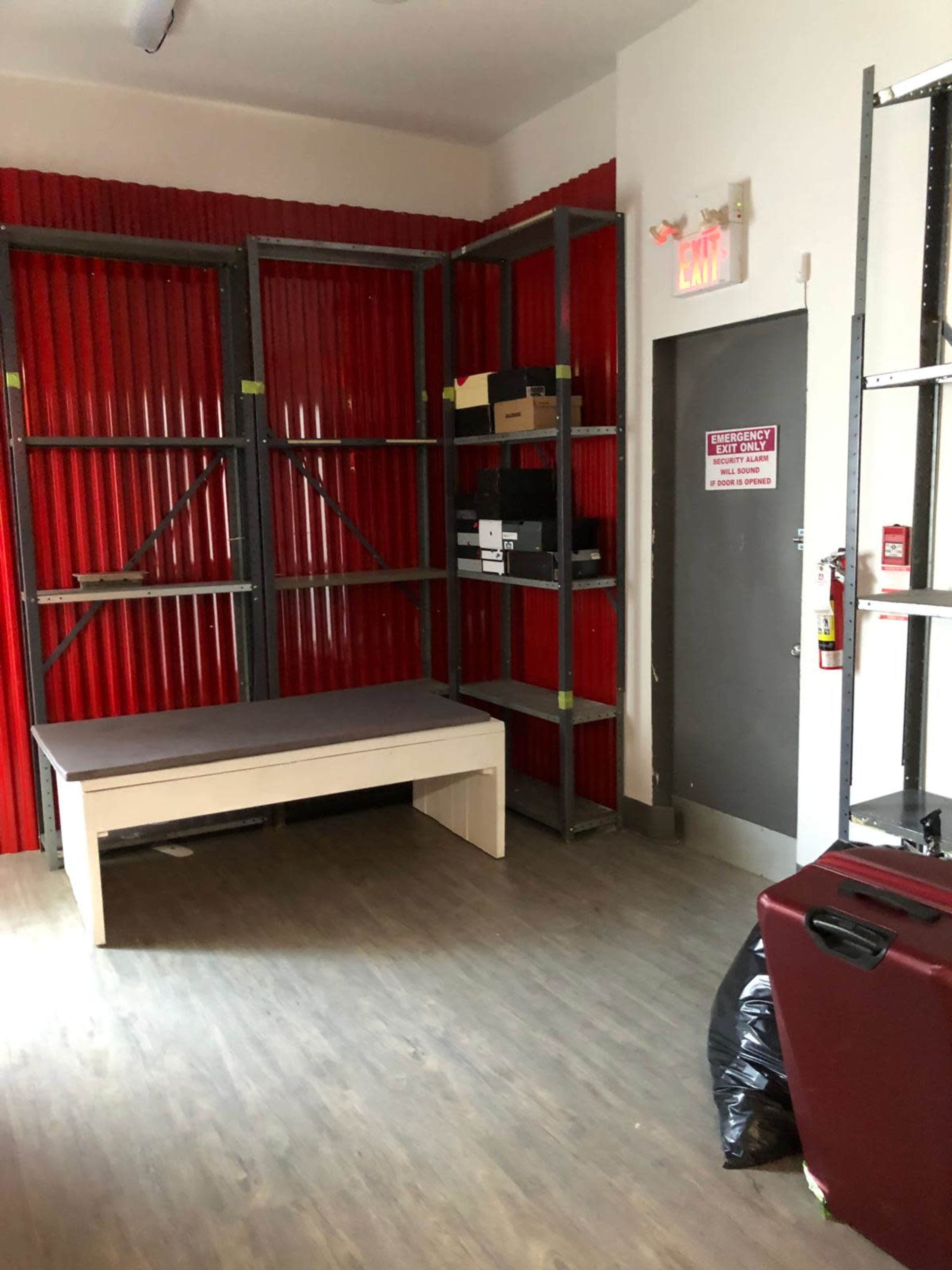 The image shows a storage room with red corrugated walls, metal shelving units, a wooden table, and various boxes stacked on the shelves.