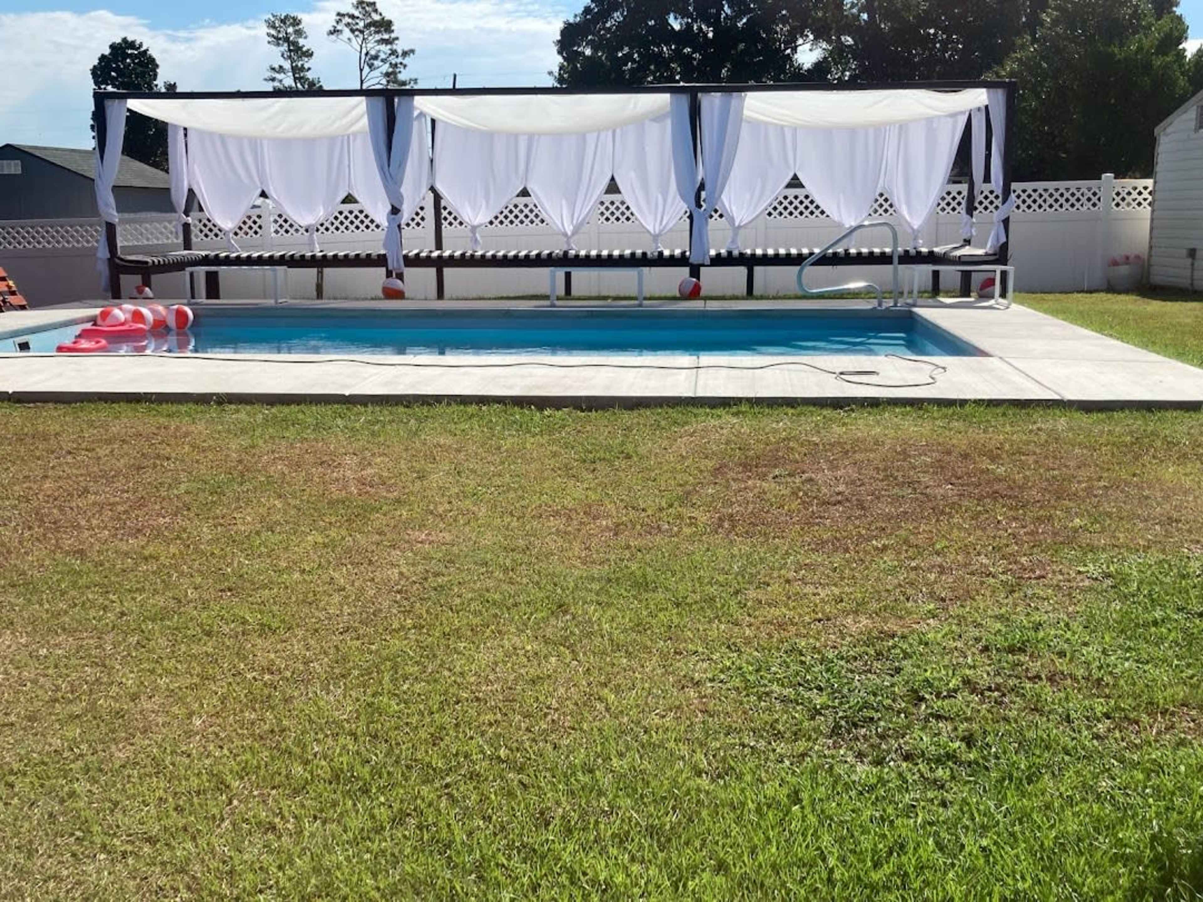 A rectangular swimming pool is surrounded by a grassy area and features a shaded seating area with white drapes.