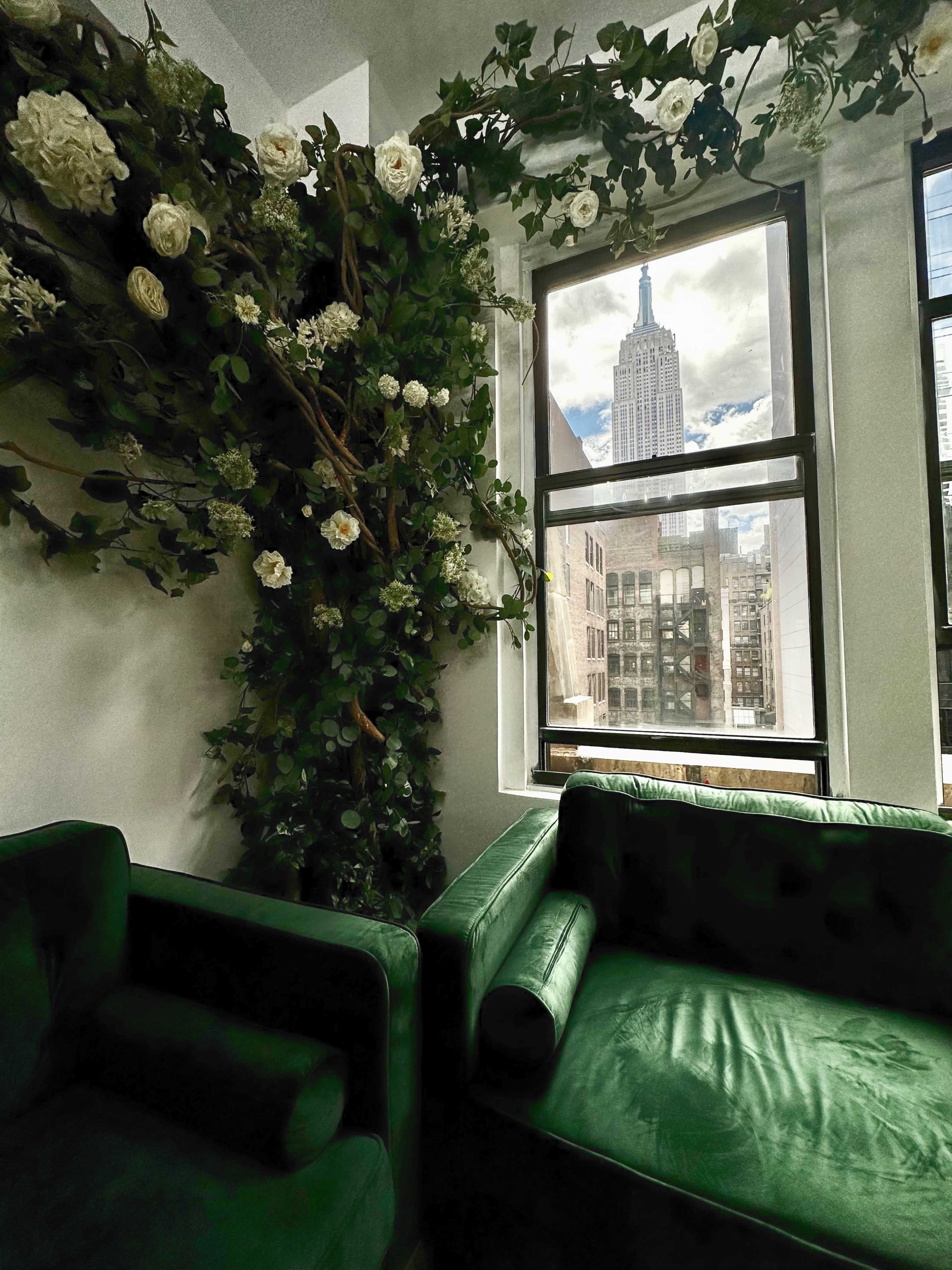 A view from inside a room featuring green couches and a window displaying the Empire State Building amidst a backdrop of clouds and foliage.