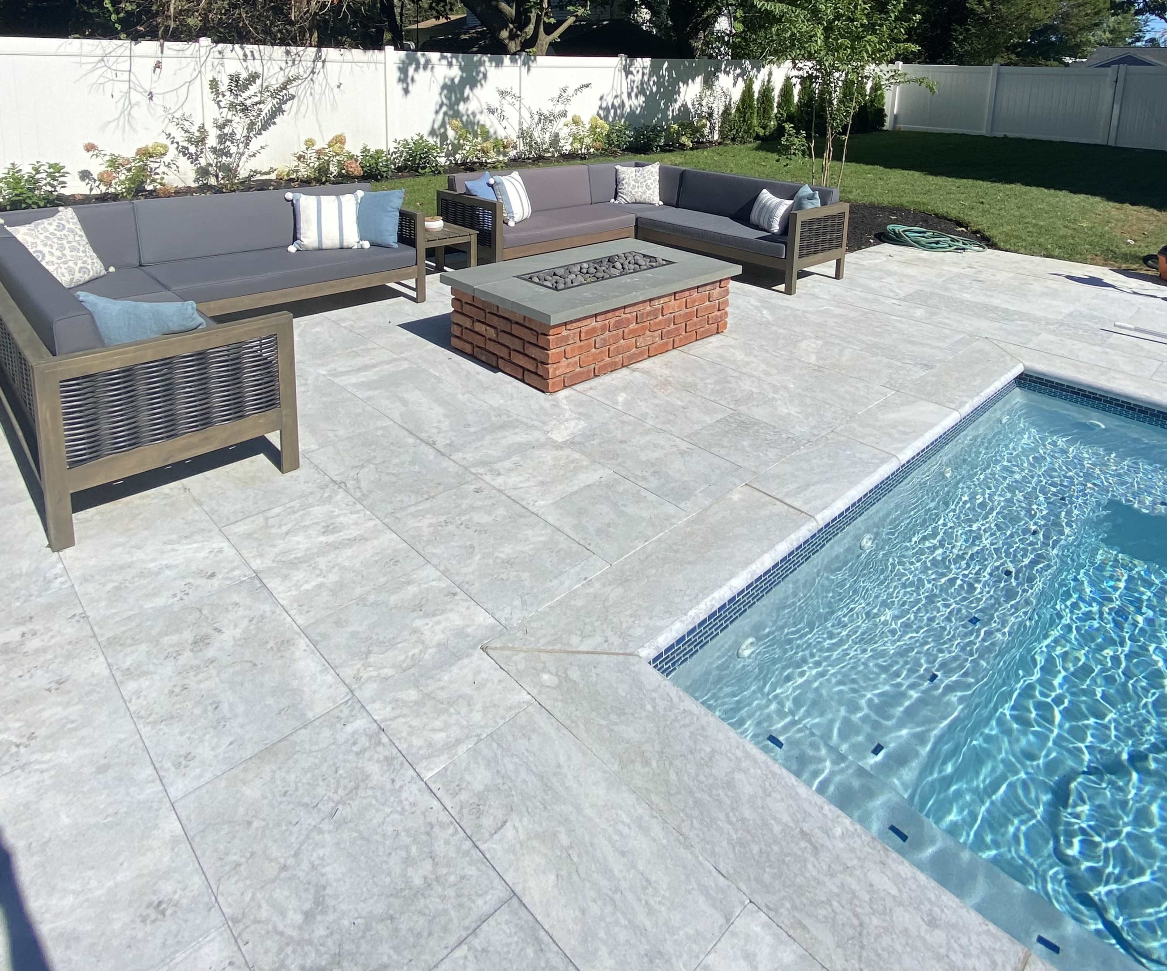 A modern backyard patio with gray stone flooring, two sectional sofas, and a brick fire pit near a swimming pool.