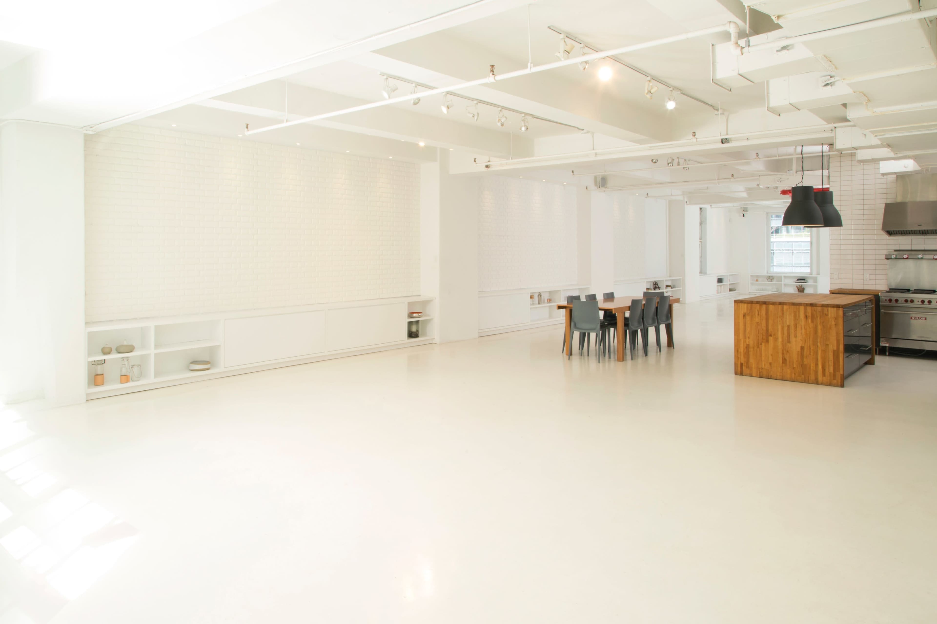 The image shows a spacious, minimalist kitchen and dining area with white walls, a large wooden island, and a simple dining table surrounded by chairs.