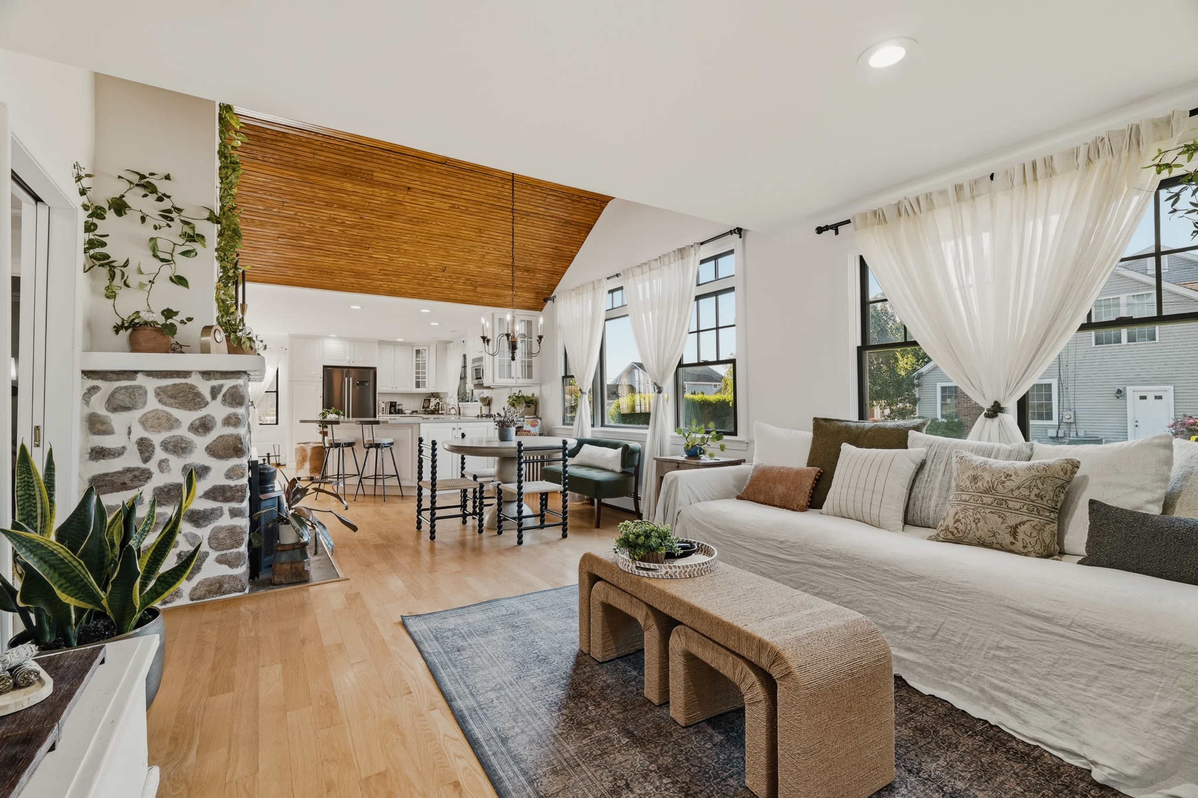 The image shows a spacious living room with large windows, a stone fireplace, and a cozy seating area adorned with various cushions and plants.