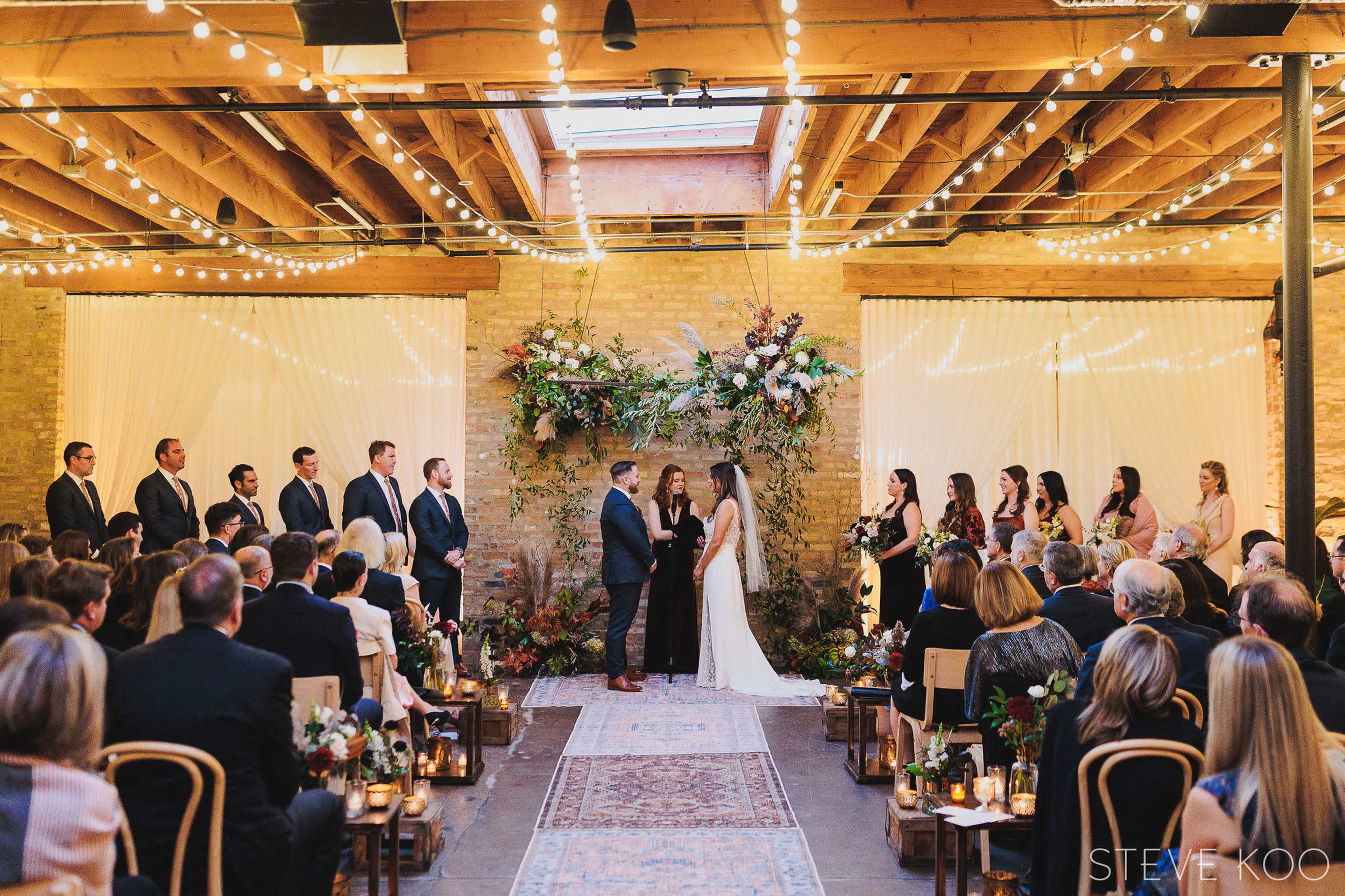 A couple stands in front of a floral arch during their wedding ceremony, surrounded by guests seated in rows.