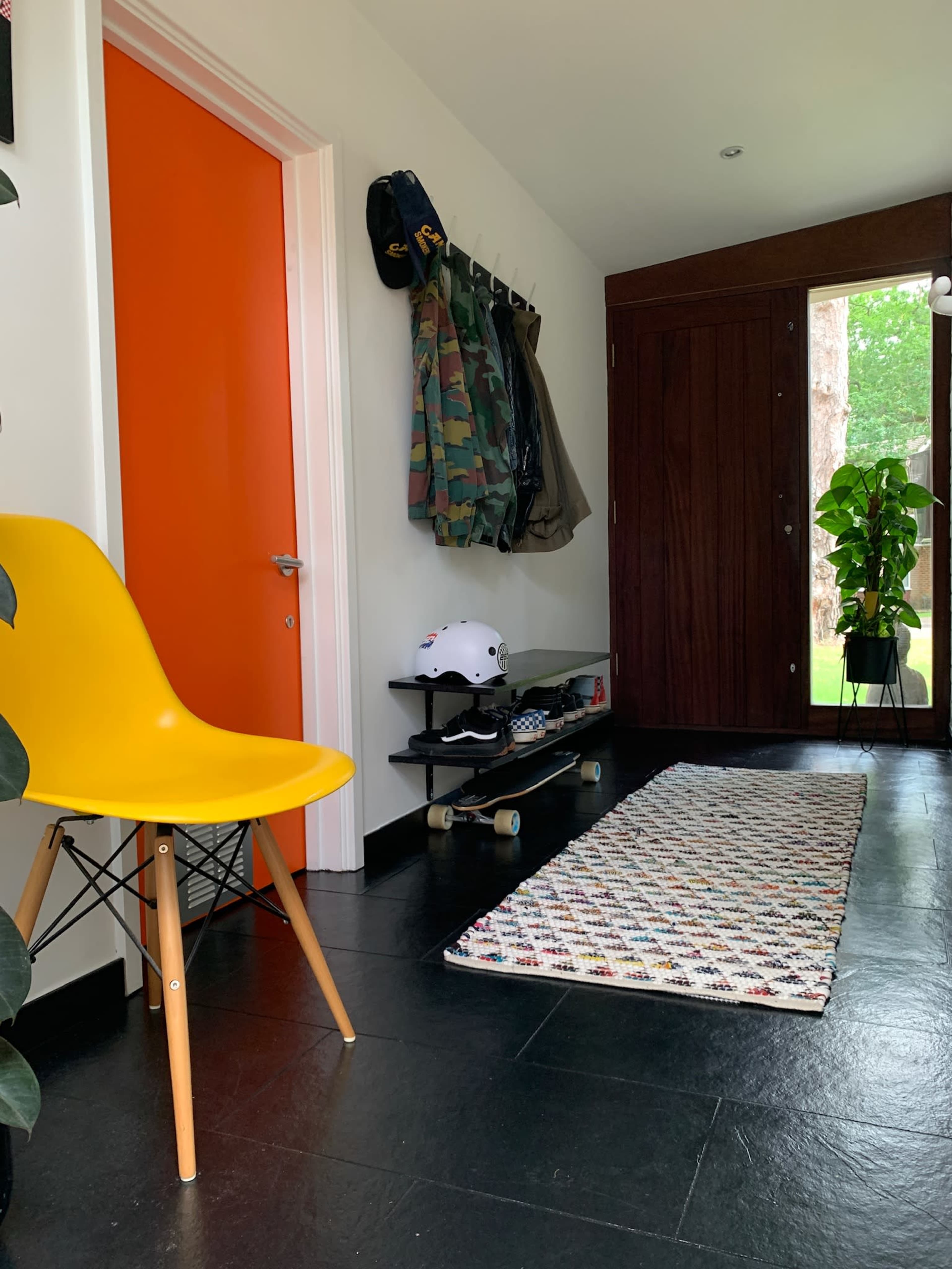 A bright orange door is flanked by a yellow chair, a skateboard, a helmet, and several jackets hanging on hooks along a wall, with a textured rug on the floor.