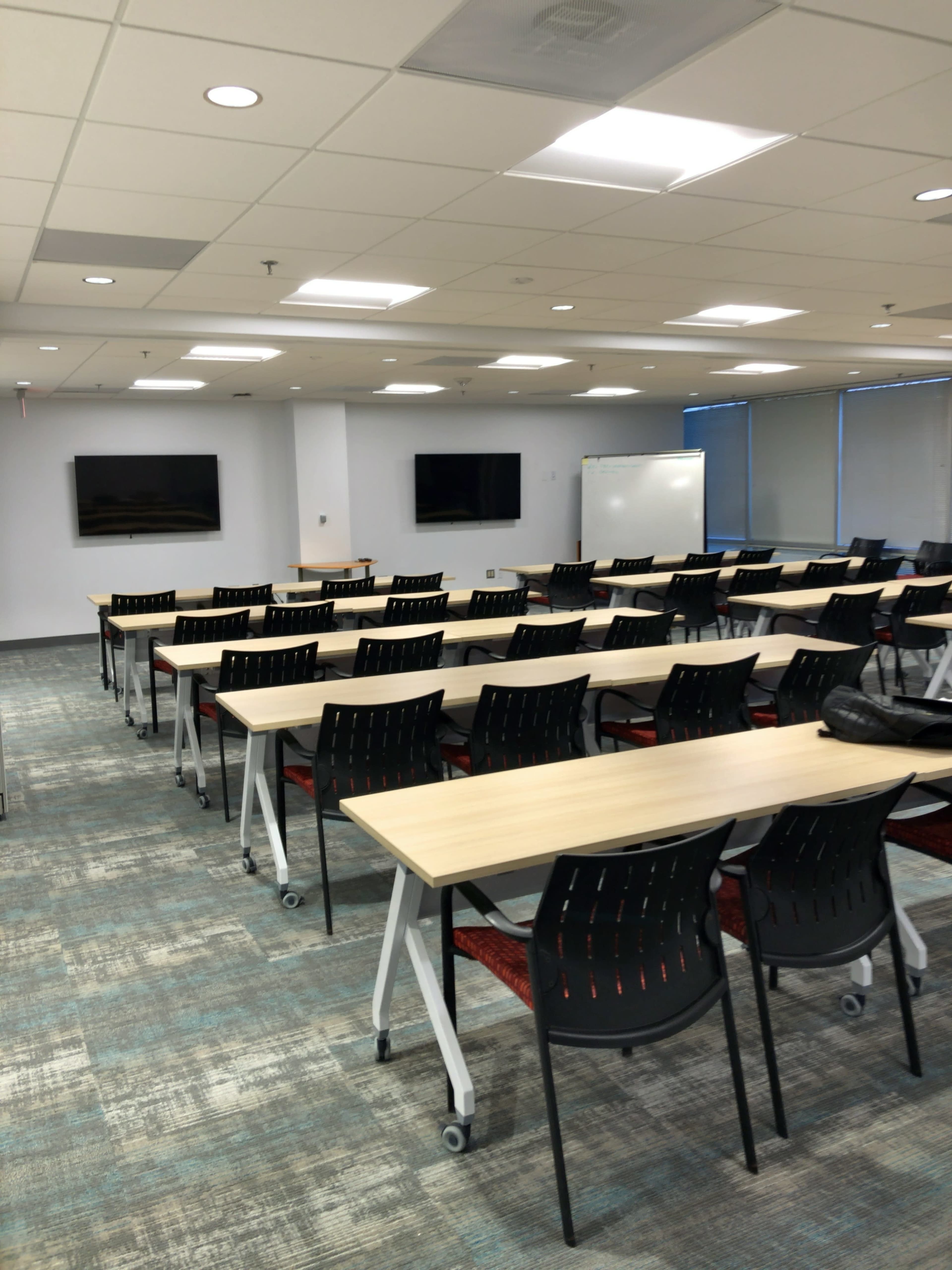 A conference room is set up with multiple rows of tables and chairs, with two screens mounted on the walls and a whiteboard in the corner.