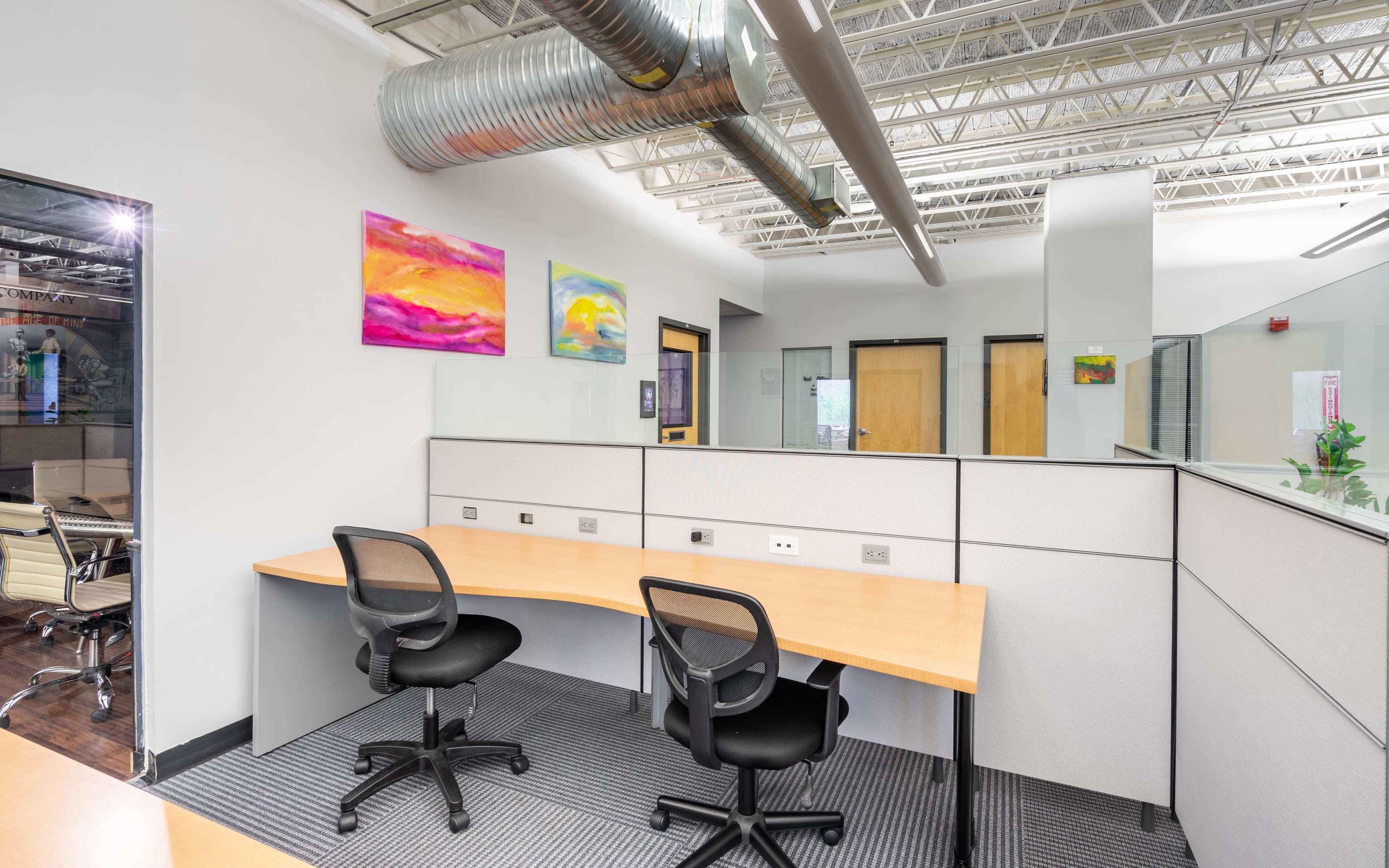 An office space with two empty desks and glass partitions, featuring colorful artwork on the walls.