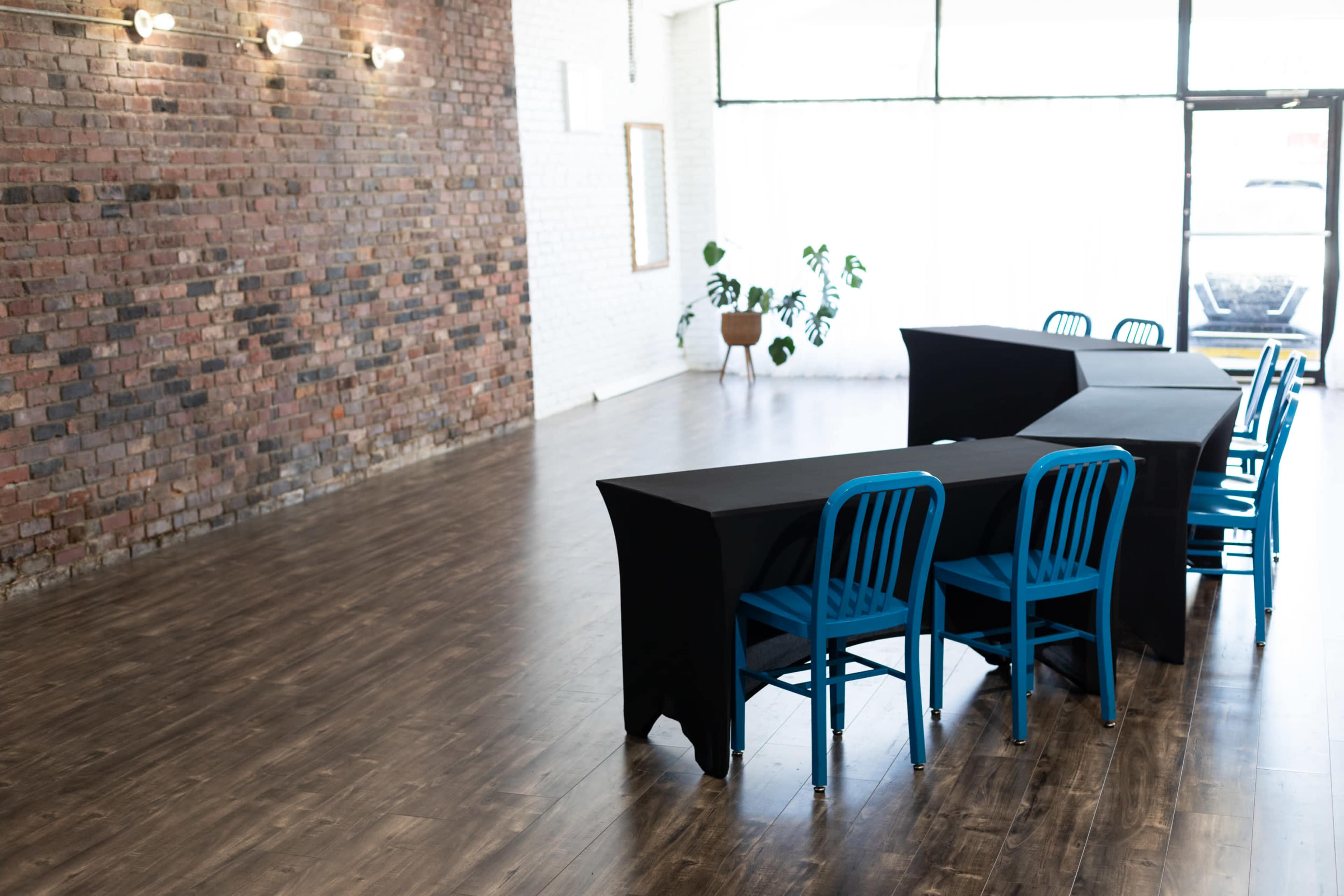 A room features a brick wall with two rectangular tables covered in black tablecloths and several blue chairs arranged nearby.