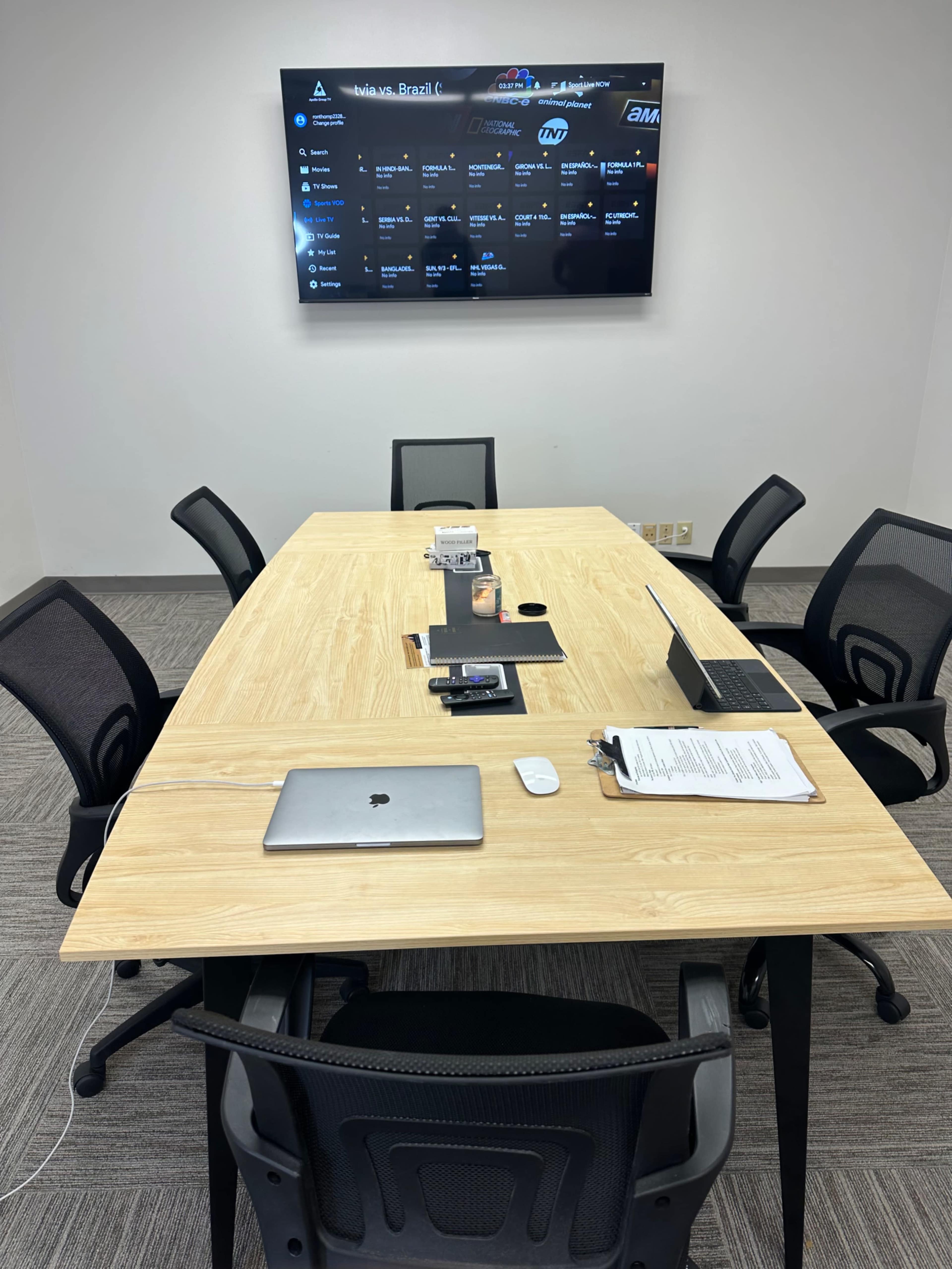 A conference room features a wooden table surrounded by black chairs, with a large screen displaying data on the wall and various electronic devices placed on the table.