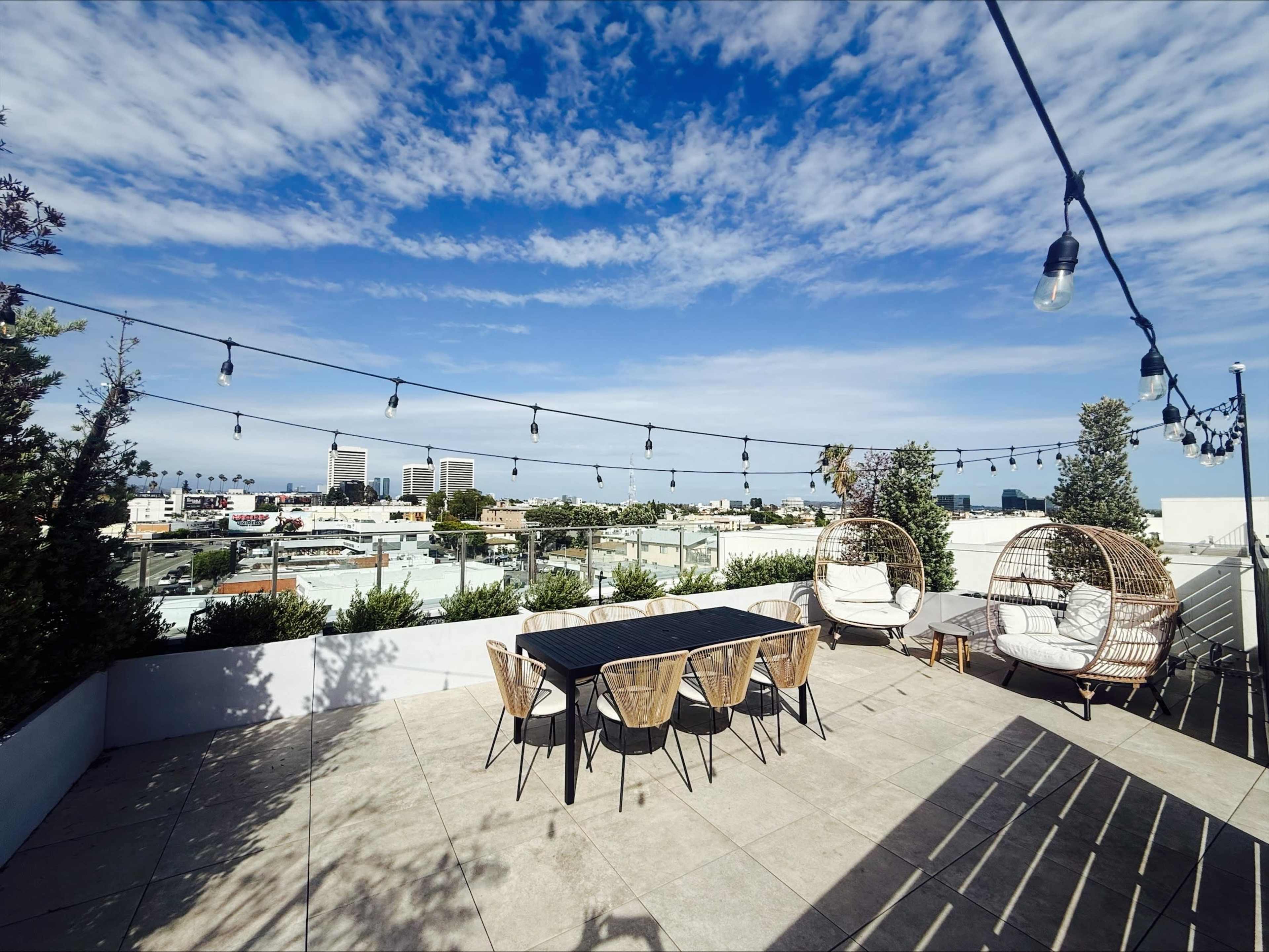 The image shows a rooftop terrace featuring a dining table with chairs, two wicker lounge chairs, and string lights overhead, overlooking a cityscape under a partly cloudy sky.