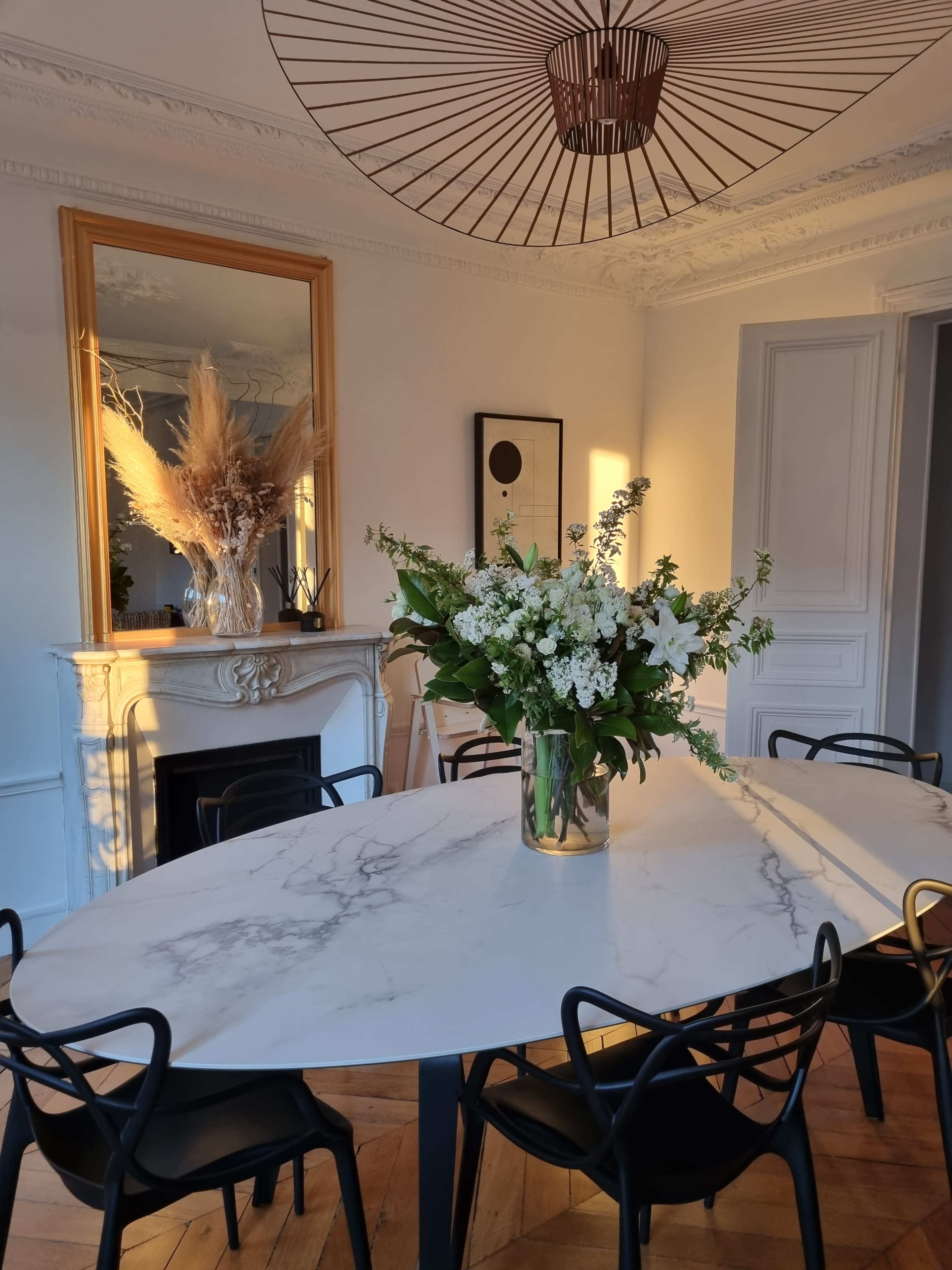A marble dining table with a floral centerpiece is set in a well-lit room featuring a large mirror and decorative accents.