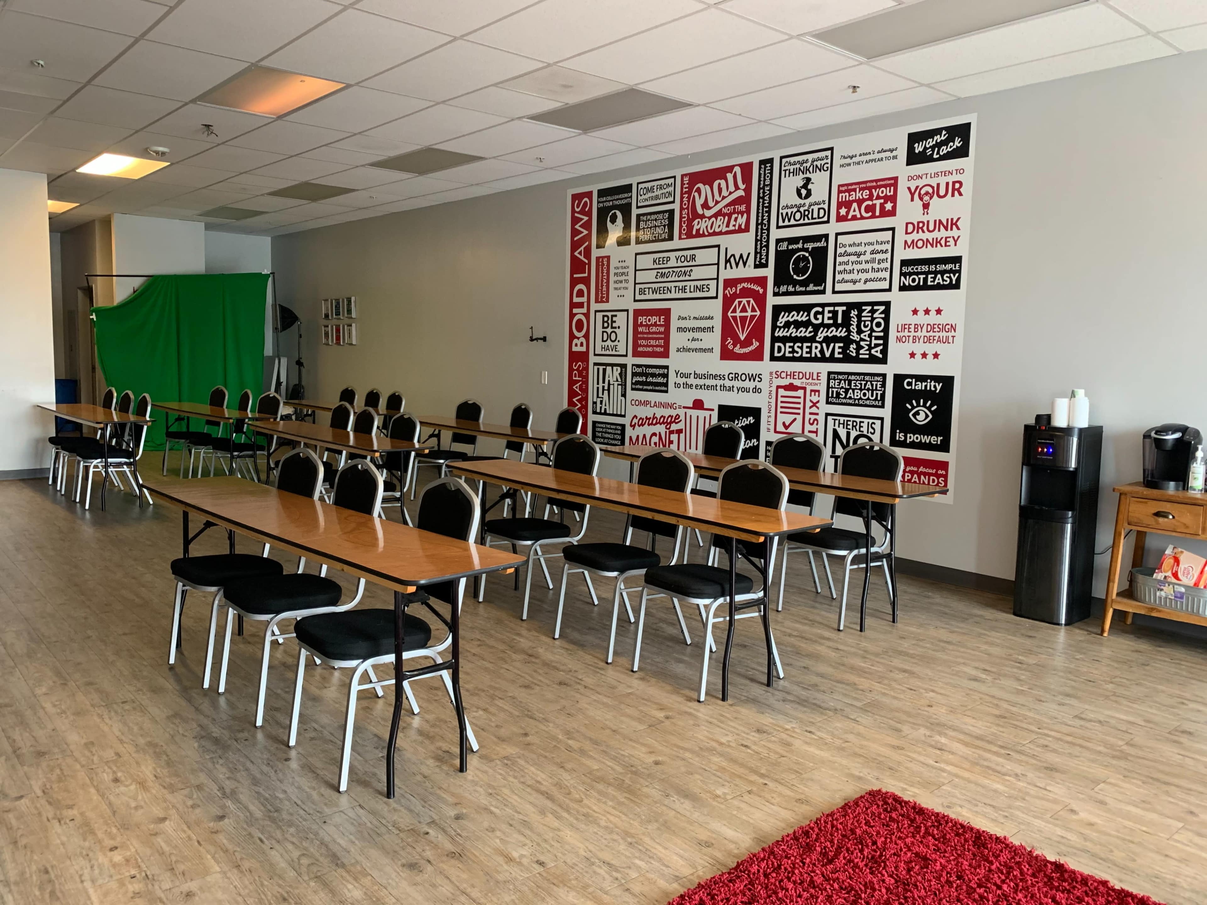 A spacious room features rows of tables and chairs against a wall adorned with various motivational posters, while a green screen and beverage station are located at one end.