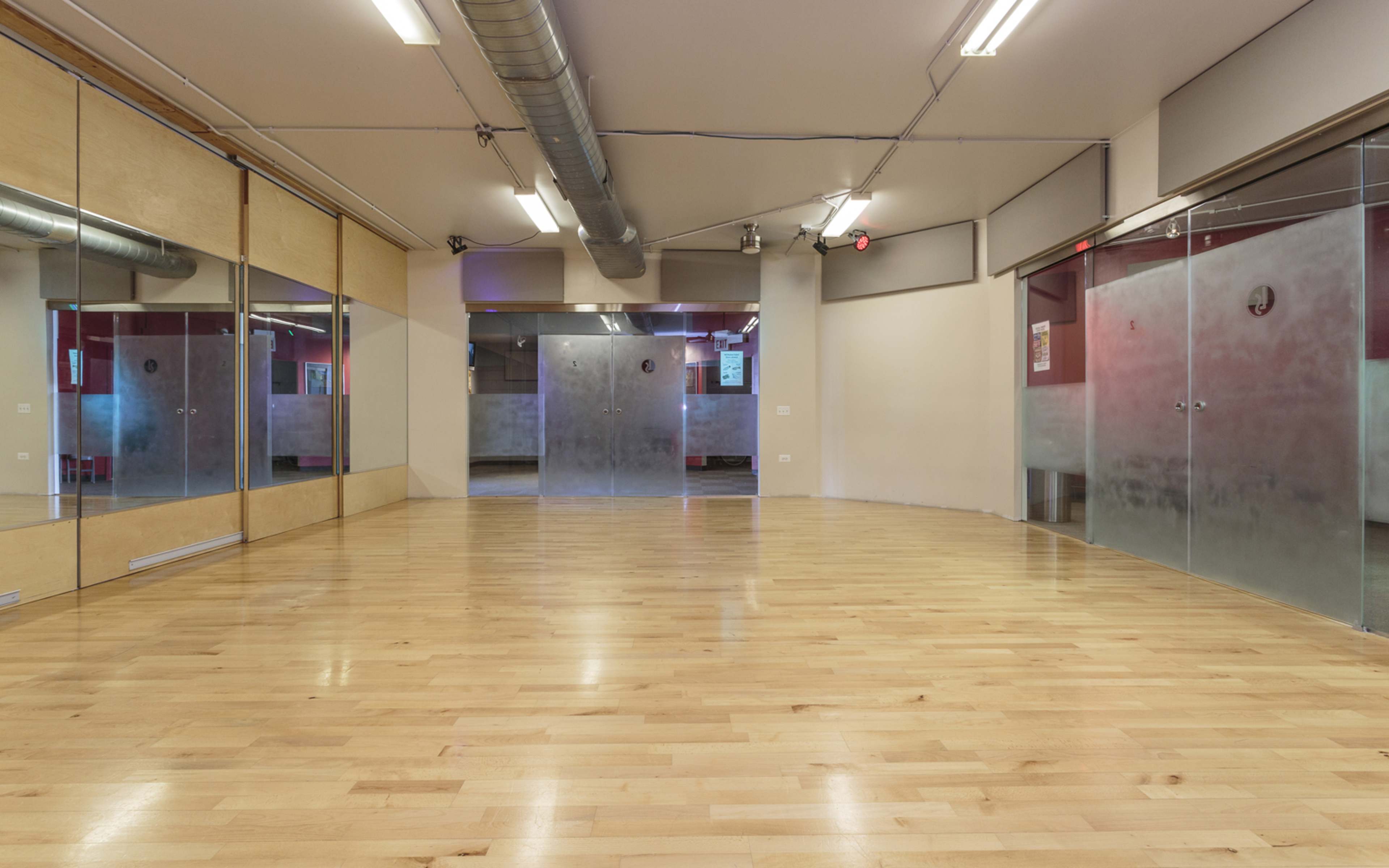 An empty studio space with wood flooring and mirrored walls, featuring glass doors at one end.
