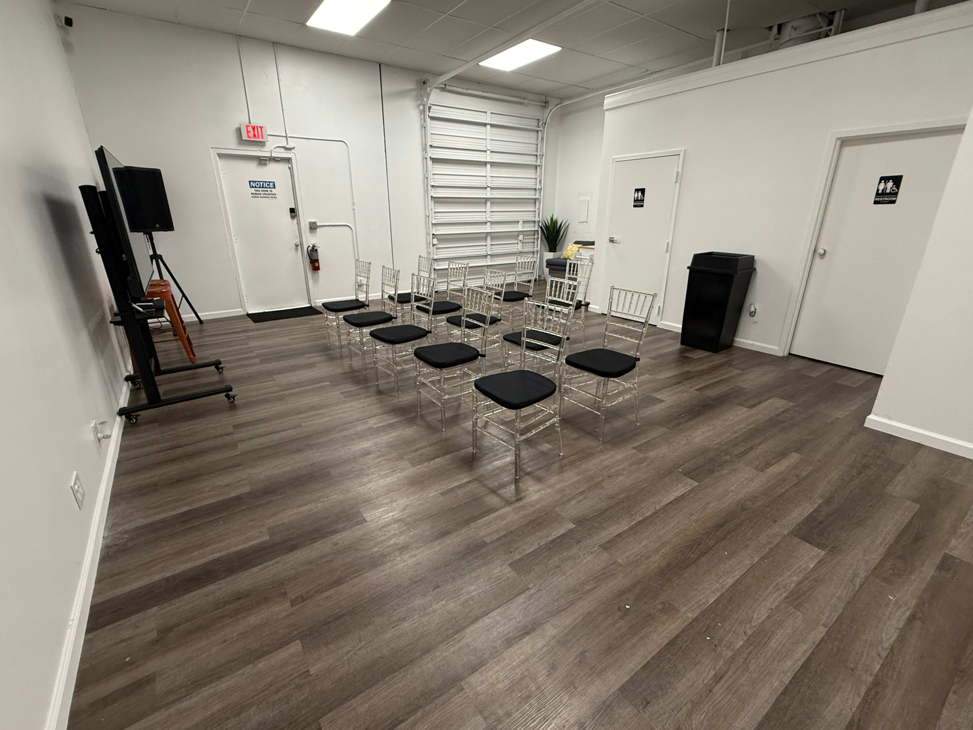 The image shows a sparsely furnished room with a row of clear acrylic chairs arranged in front of a television, and two doors labeled for restrooms.