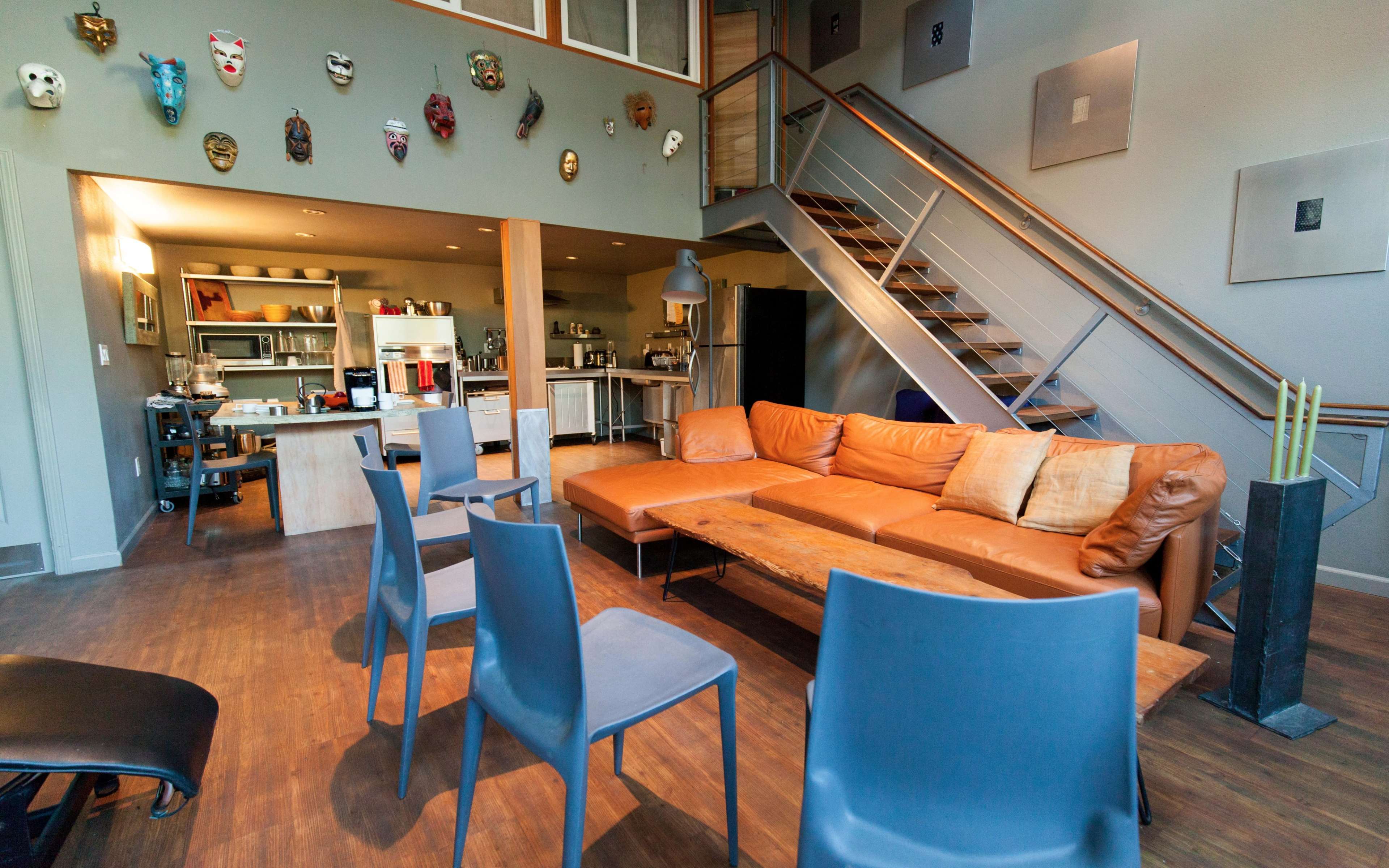 A modern living space with a brown leather sectional sofa, several gray chairs, and a staircase leading to an upper level, adorned with various masks on the walls.