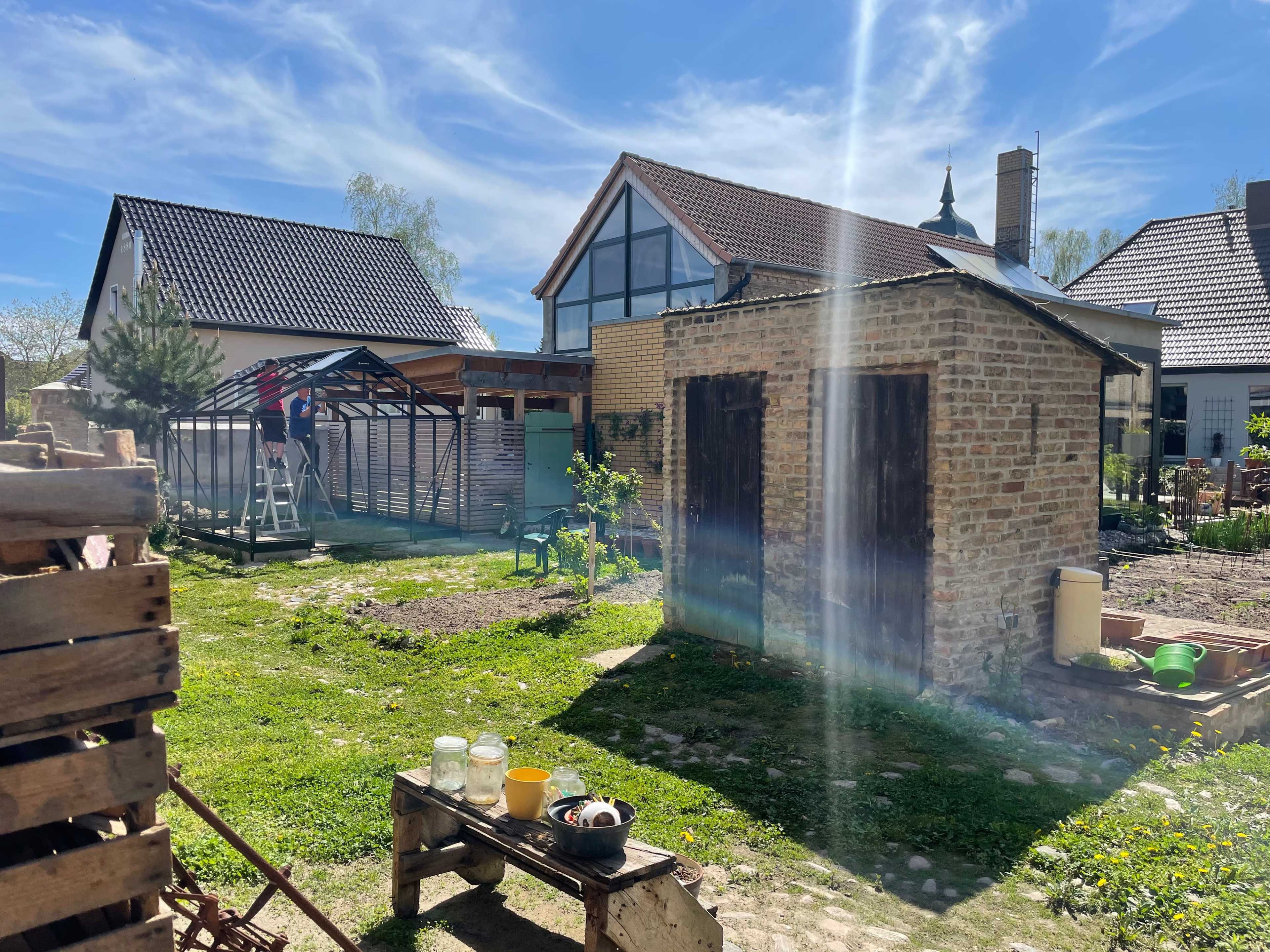 A backyard scene features a small stone shed, a wooden structure under construction, and a nearby house, all set against a clear blue sky.