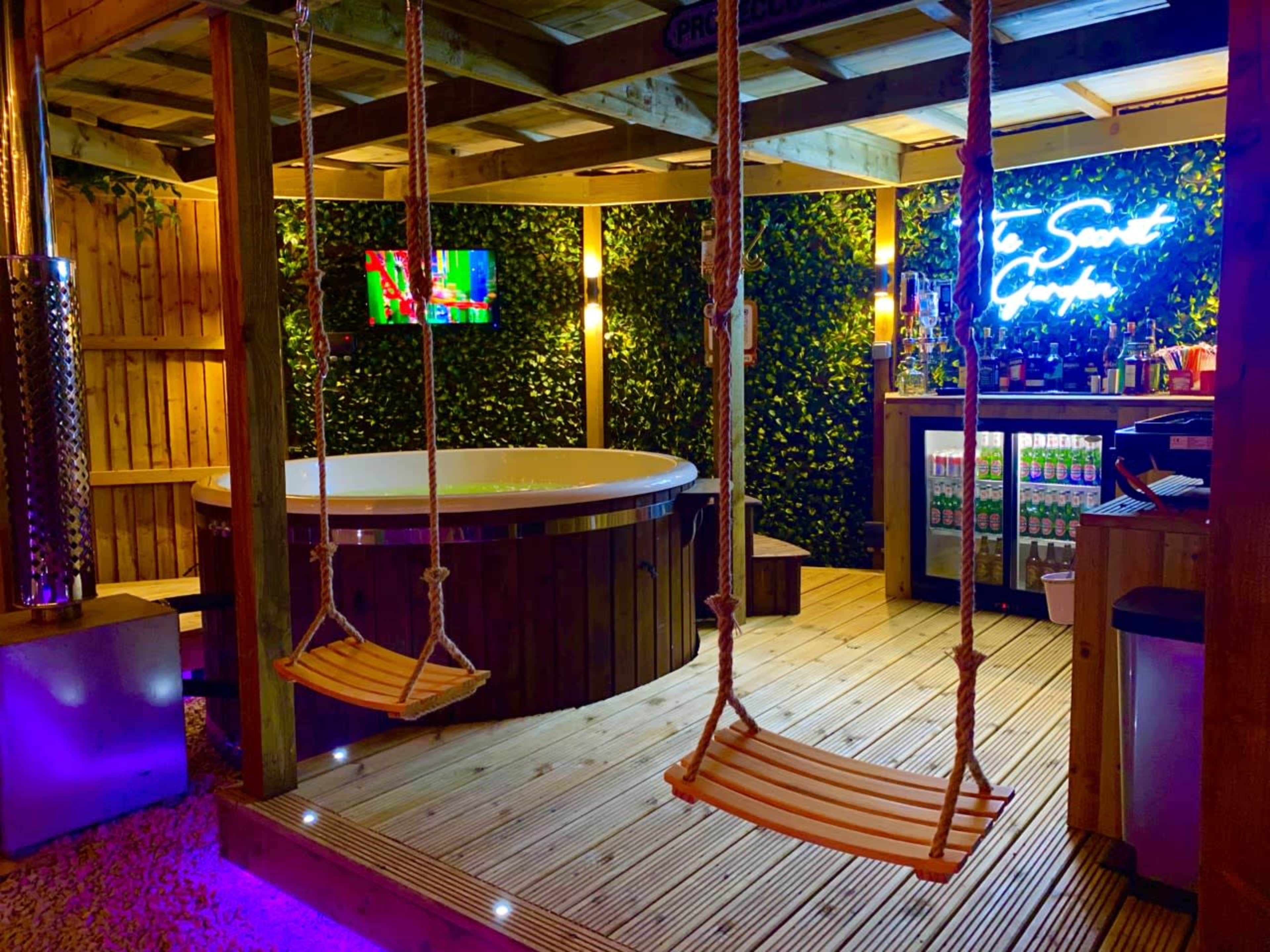 A wooden deck area with a hot tub, two swings hanging from a pergola, and a bar with an illuminated sign in a lush, green setting.