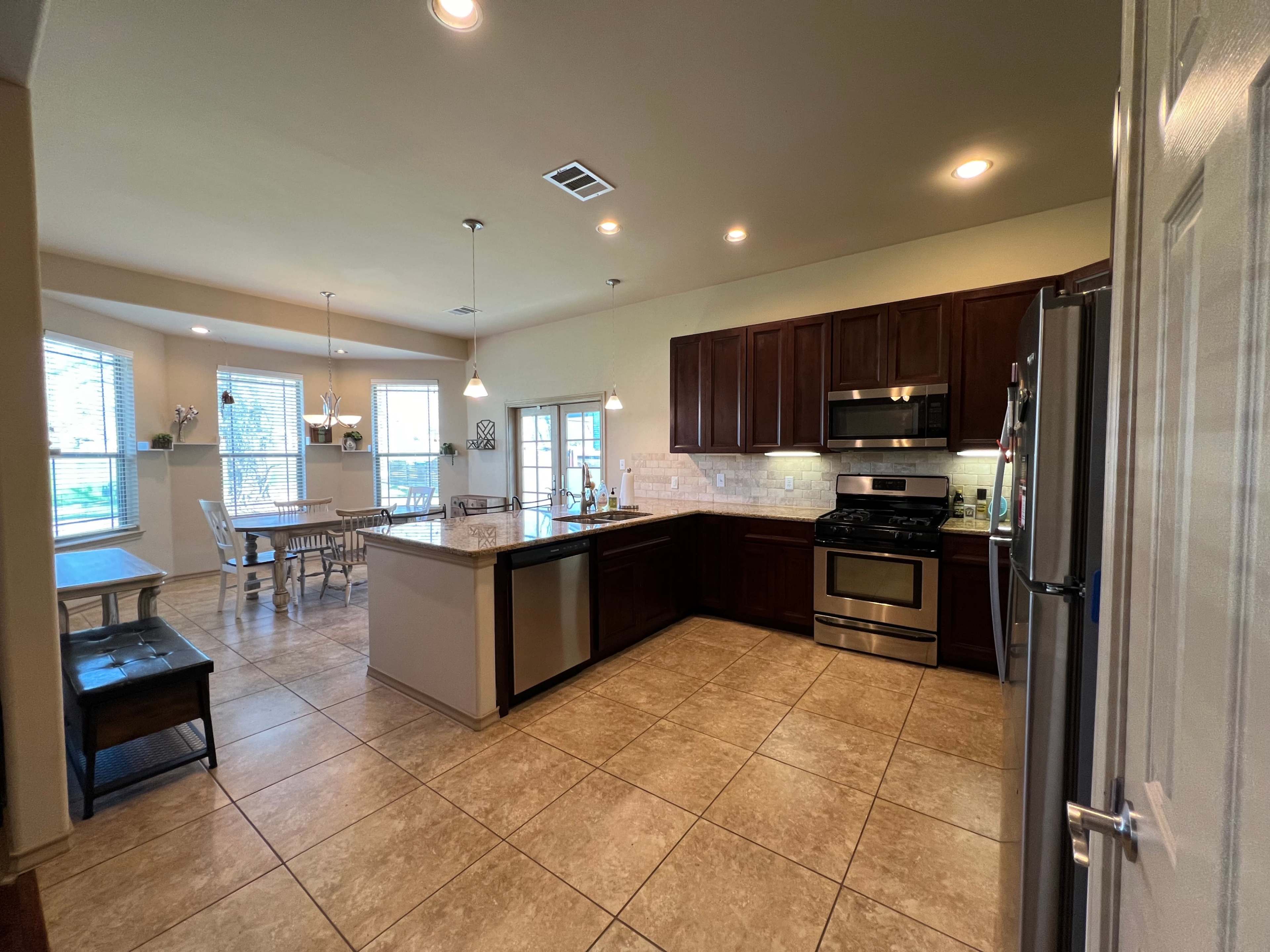 A modern kitchen with dark wooden cabinets, stainless steel appliances, and a spacious dining area featuring a table and chairs beside large windows.