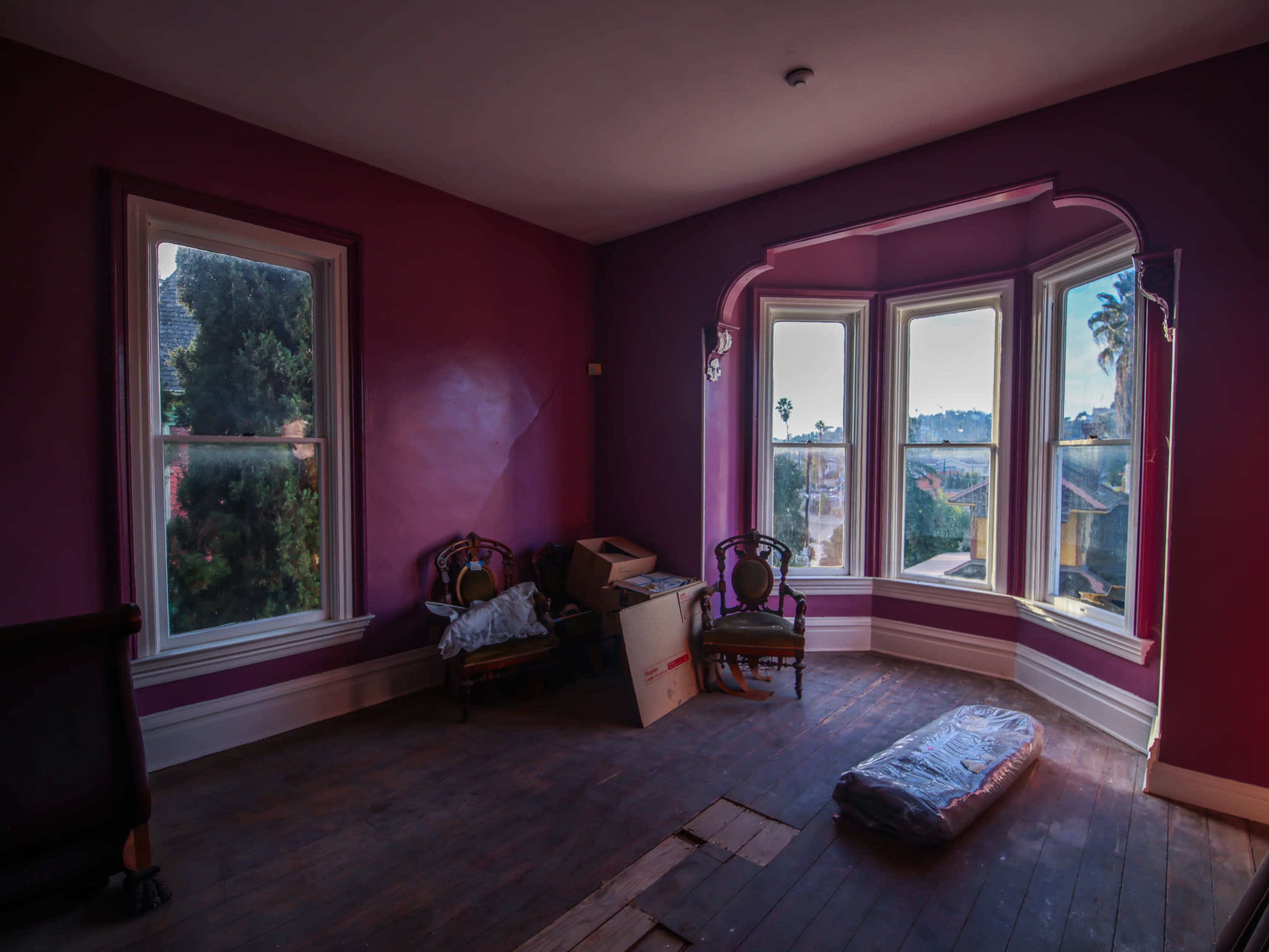 The room features purple walls, two windows with a view outside, two antique chairs, a cardboard box, and a rolled-up rug on the wooden floor.