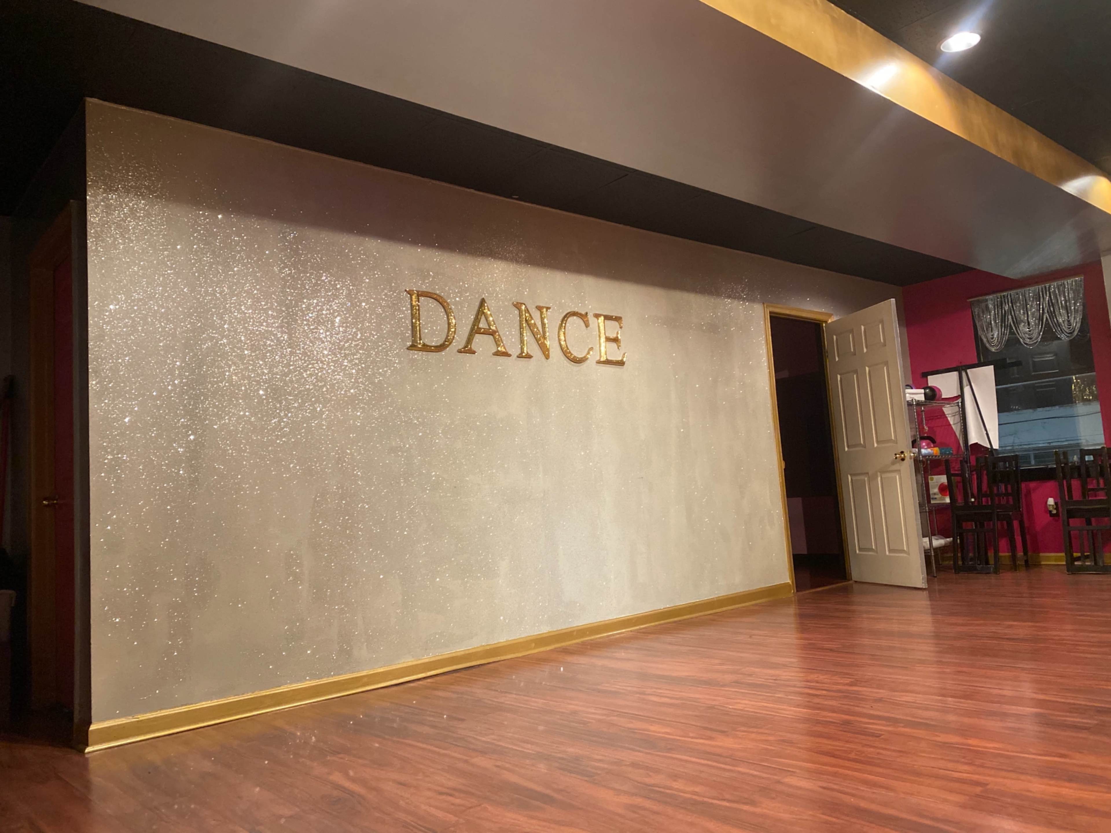 A dance studio with a shimmering wall that has the word "DANCE" prominently displayed in gold letters, and a doorway leading to another area.