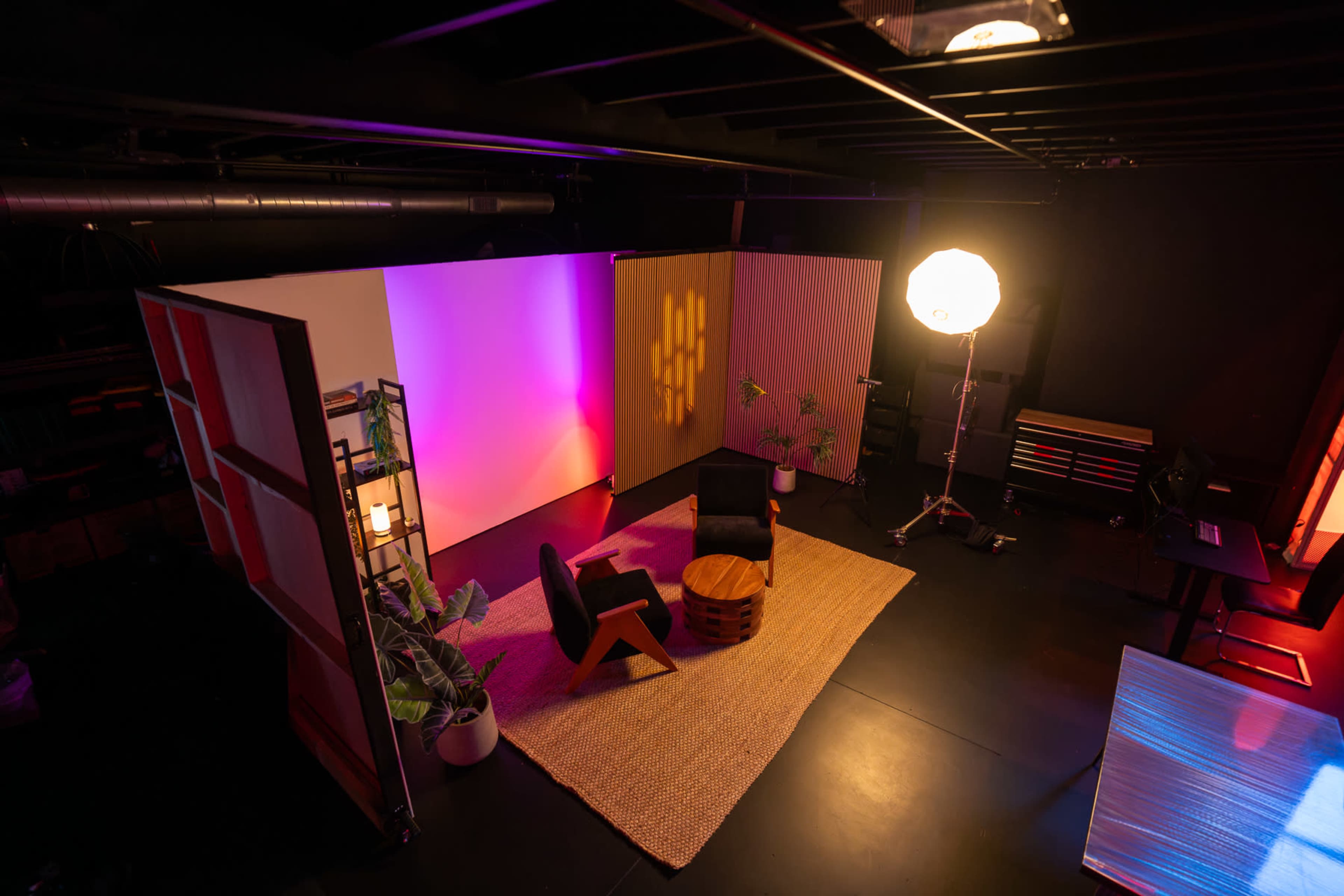 The image shows a modern studio setup featuring two chairs, a round wooden table, plants, and colorful lighting on the walls.