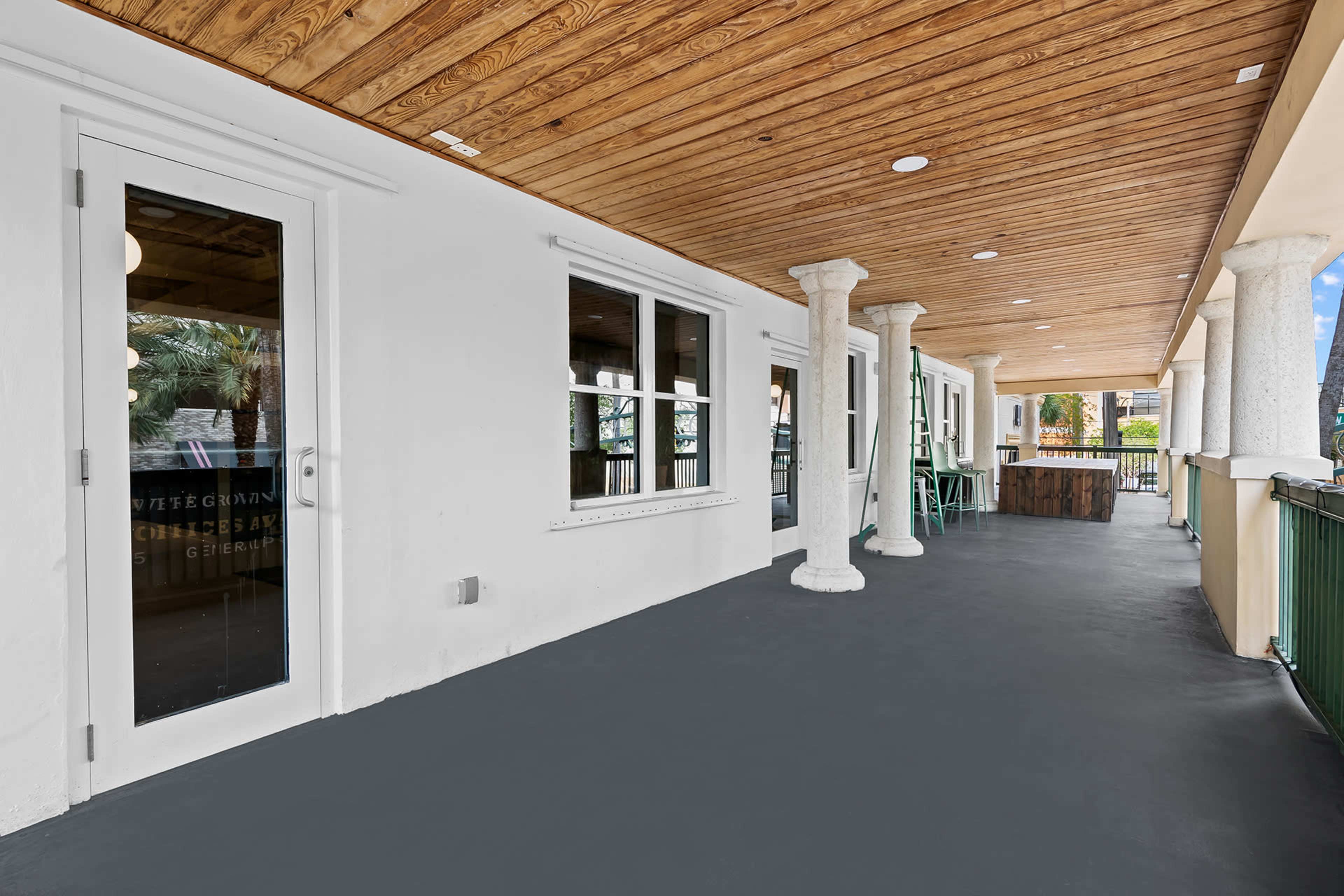 The image shows a spacious covered balcony with wooden ceiling beams, white walls, and a dark gray floor.