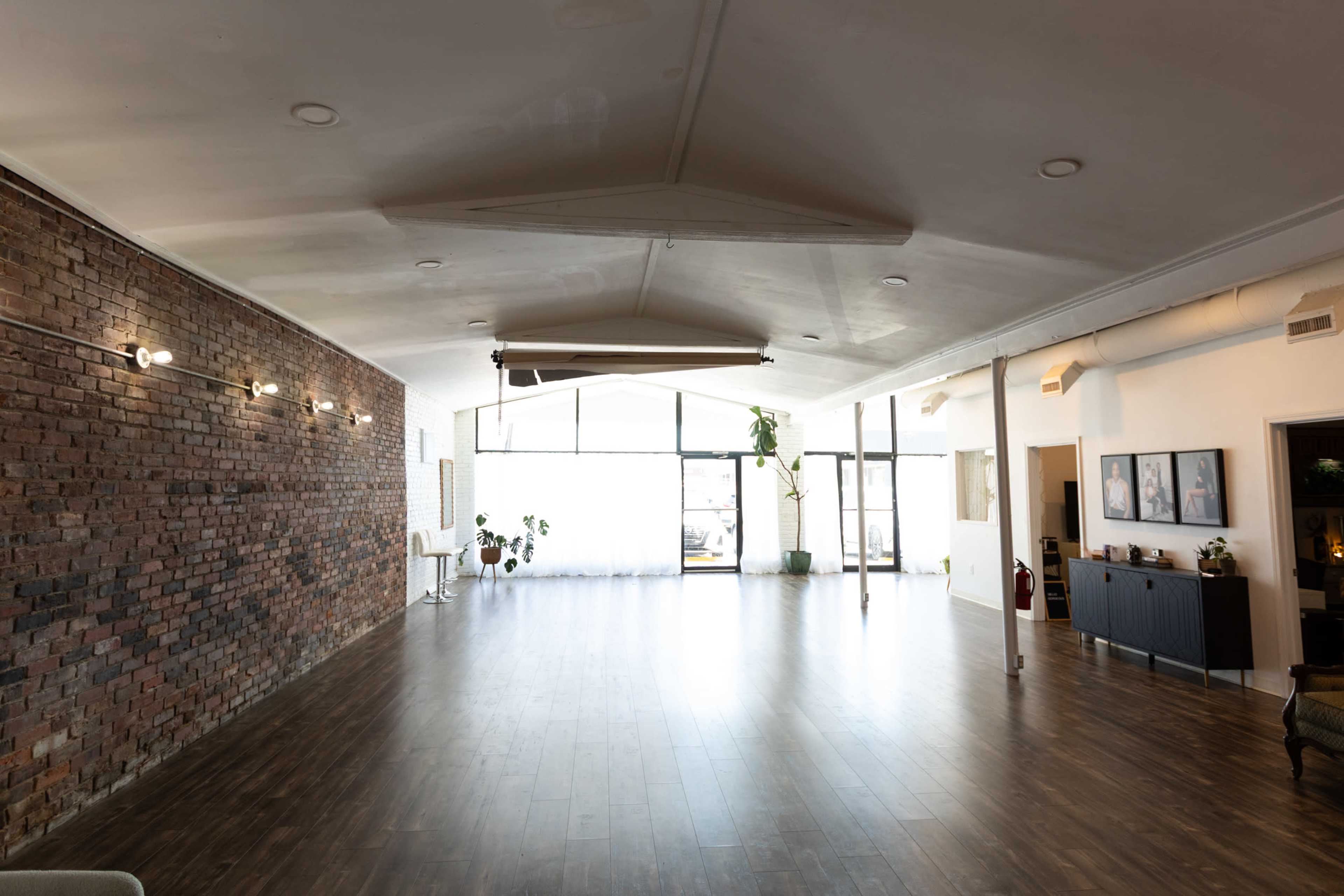 The image shows a spacious interior with a hardwood floor, brick walls, and large windows allowing natural light to fill the room.