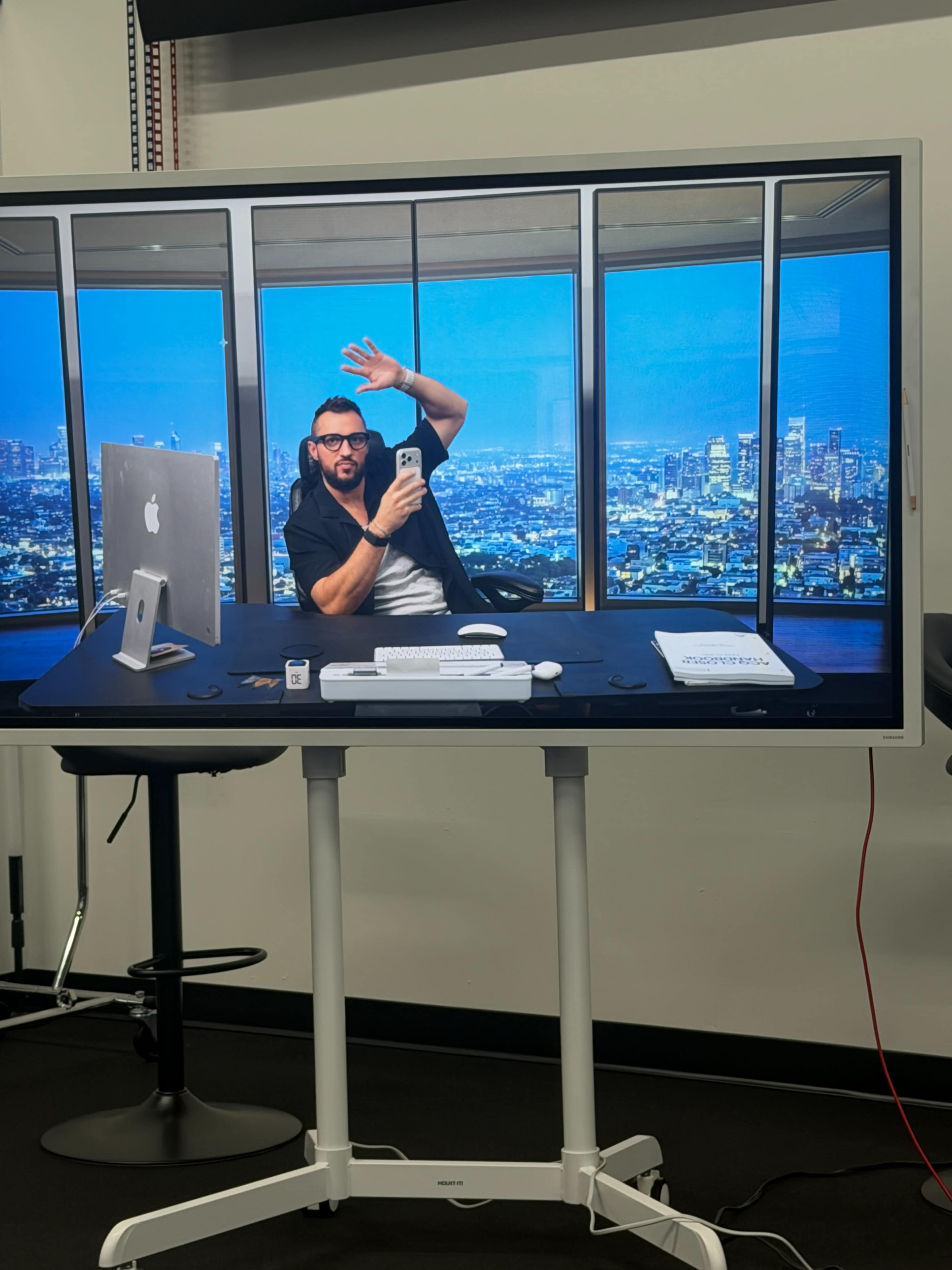 A man is sitting at a desk with an Apple computer and a smartphone, waving while surrounded by a city skyline visible through large windows in a modern office setting.