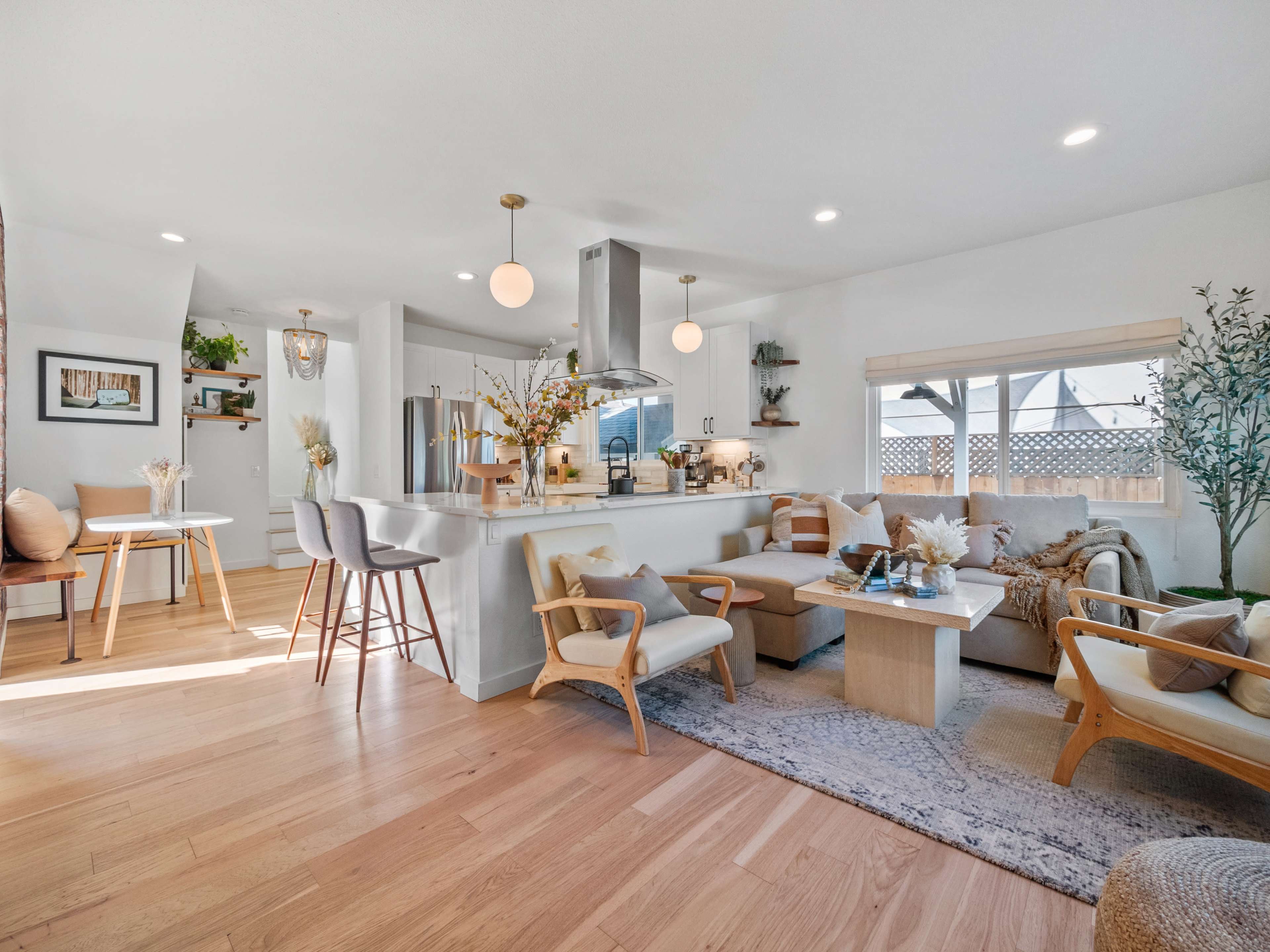 A bright, modern living space features a kitchen and dining area with wooden furnishings, a light color palette, and large windows allowing for natural light.