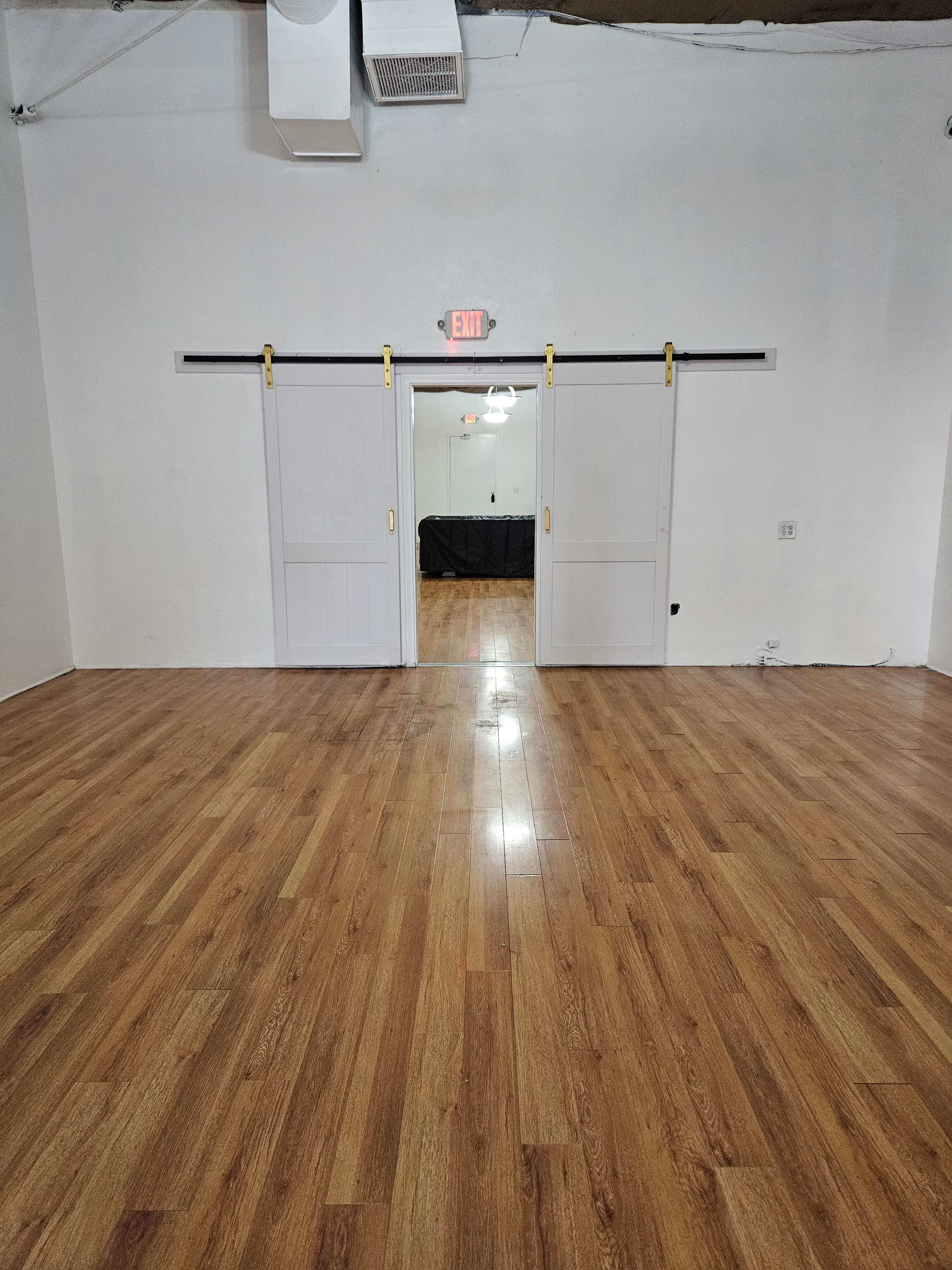 The image shows a room with light wooden flooring and two sliding barn doors in the center, leading to another room with an exit sign above.