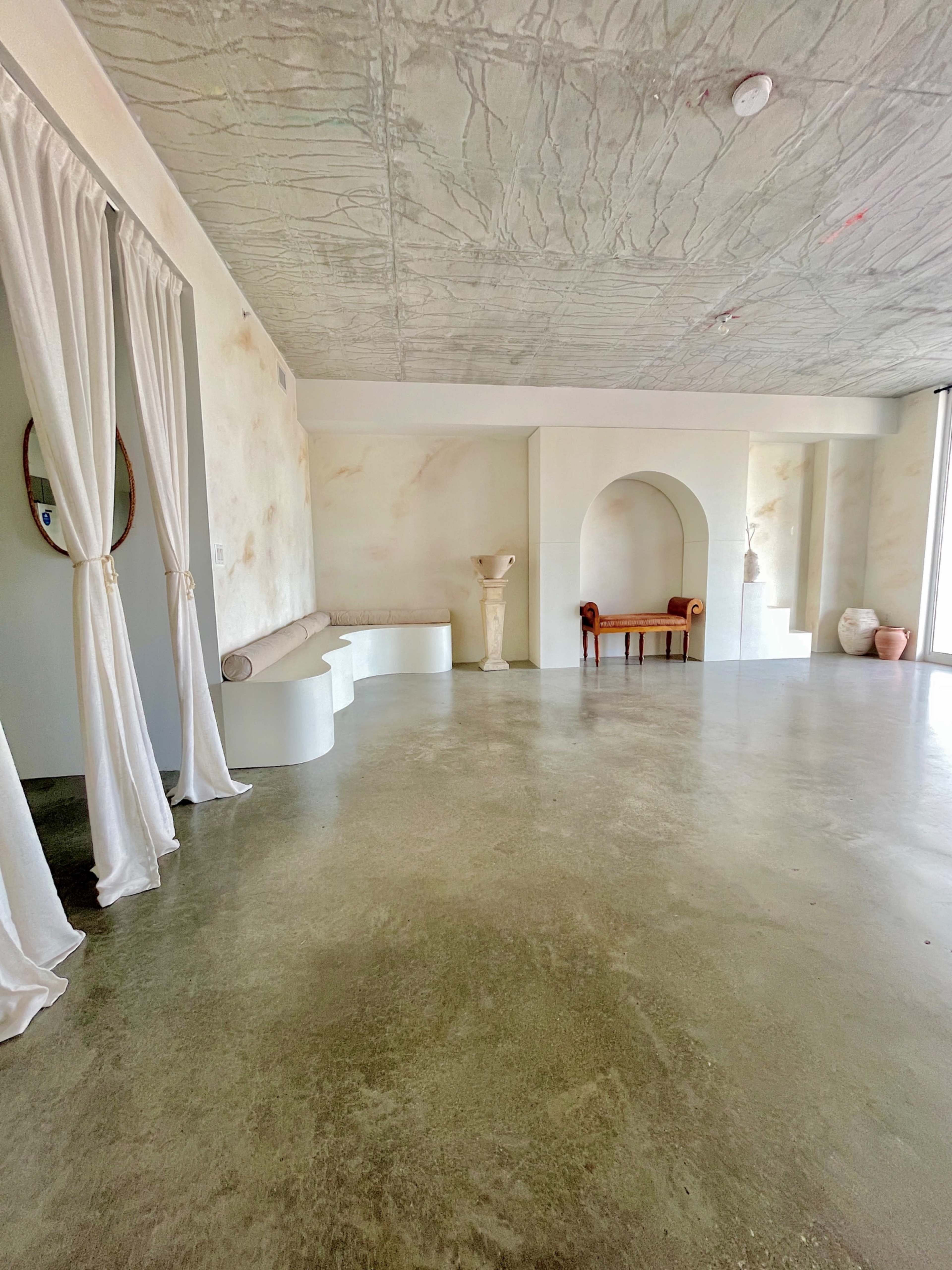 The image shows a spacious room with concrete flooring, light-colored walls, a curved bench, a sculpture, and a wooden chair against a wall arch.