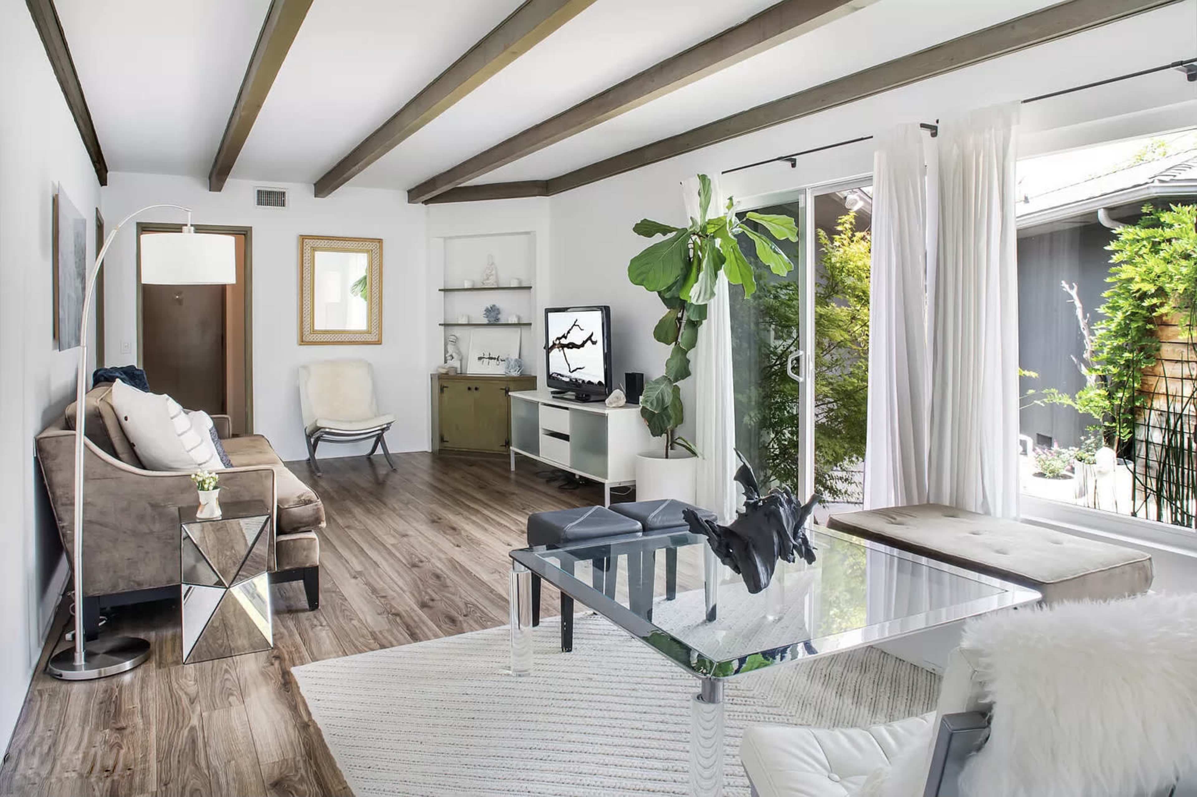A modern living room with wooden beams on the ceiling, a glass coffee table, and large windows allowing natural light to enter.