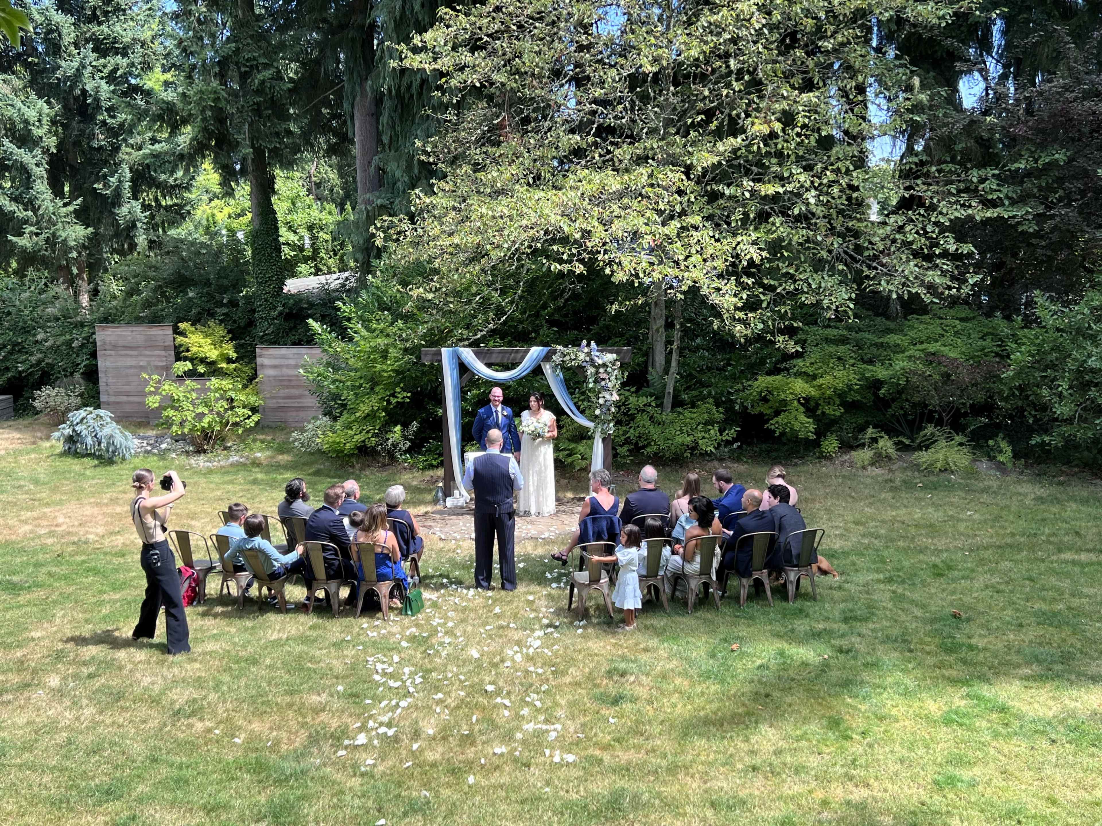 A wedding ceremony is taking place outdoors under a decorative arch, with guests seated on chairs arranged in a semi-circle on the grass.