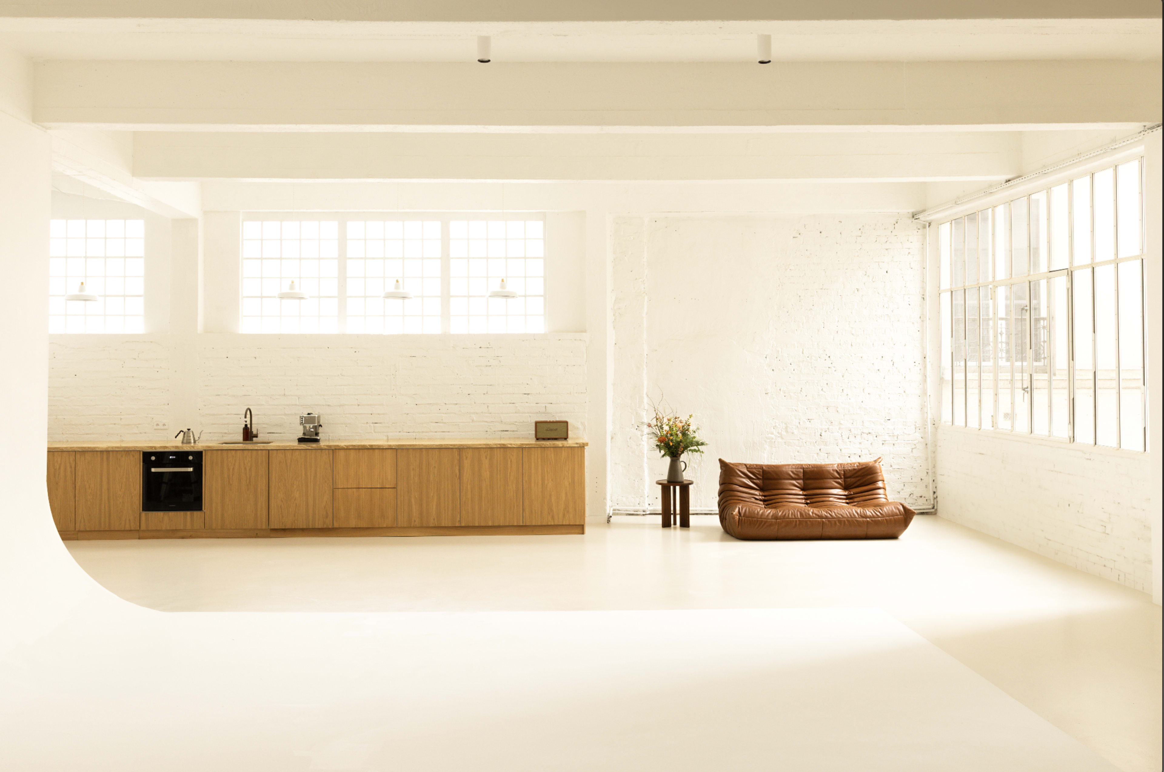 A spacious, minimalist interior with a kitchen area on one side and a brown sofa positioned near large windows on the opposite side.