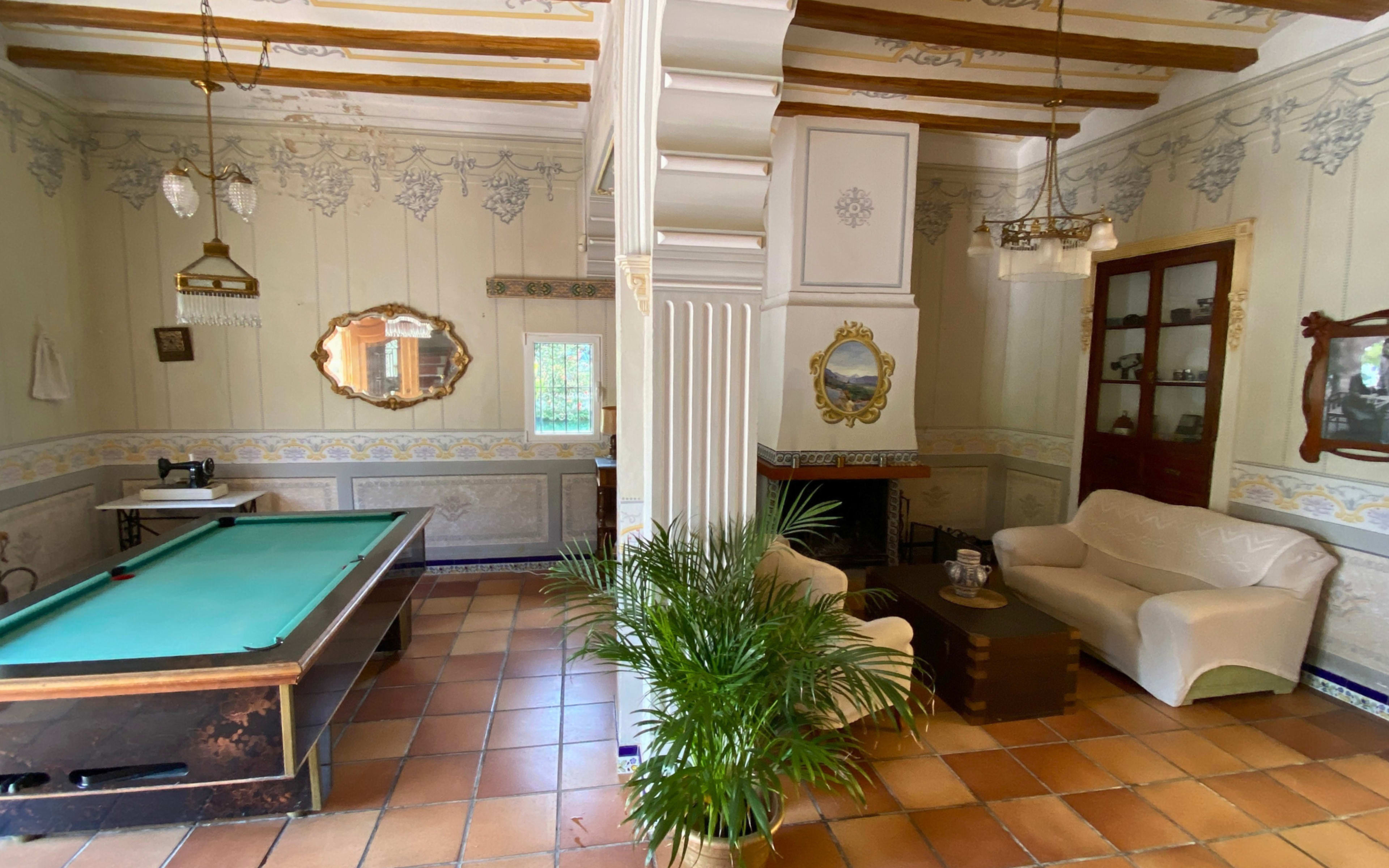 The image shows a room with a pool table on one side, and a seating area that includes a white sofa and a coffee table on the other side, with decorative wall designs and wooden beams above.