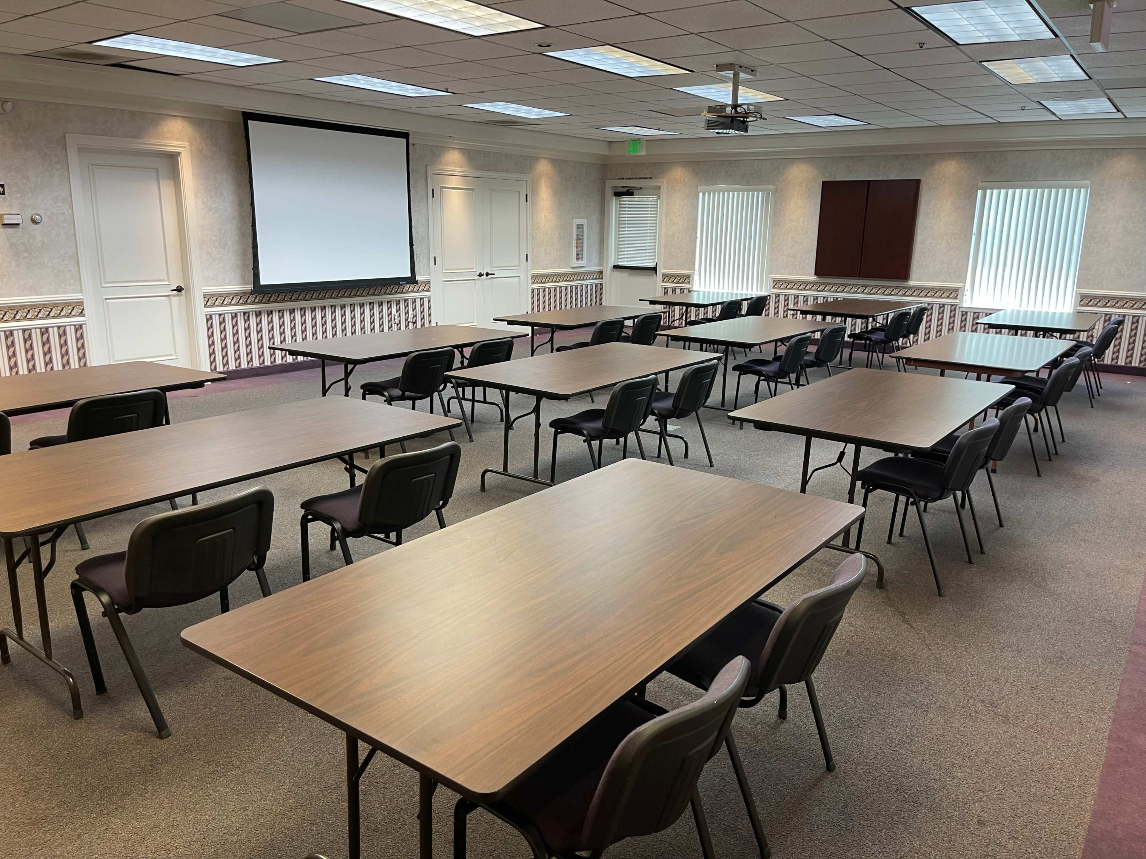 A conference room arranged with multiple tables and chairs, facing a projection screen and windows.