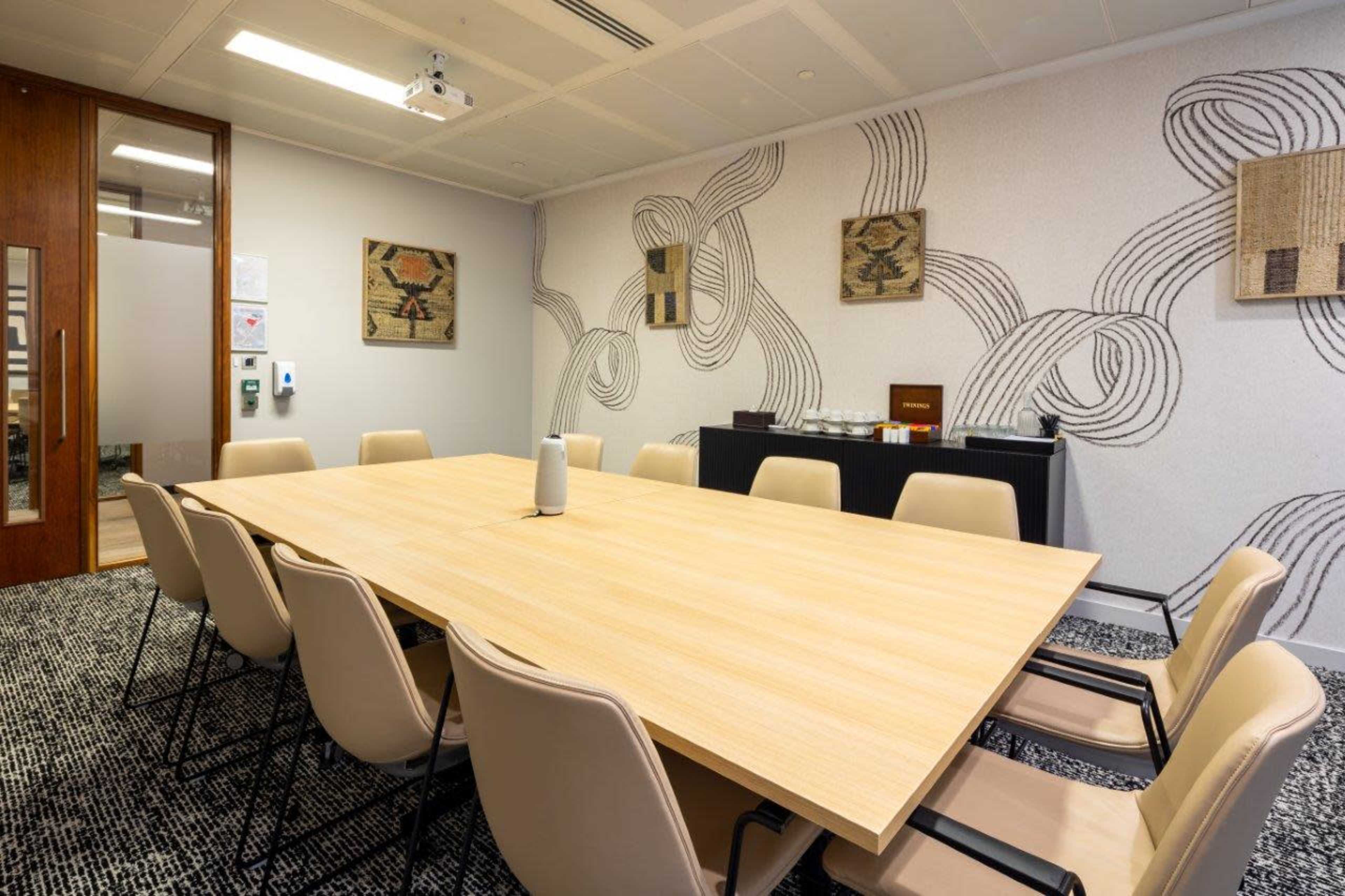 A conference room features a large wooden table surrounded by eight chairs, with patterned walls and a side table displaying refreshments.