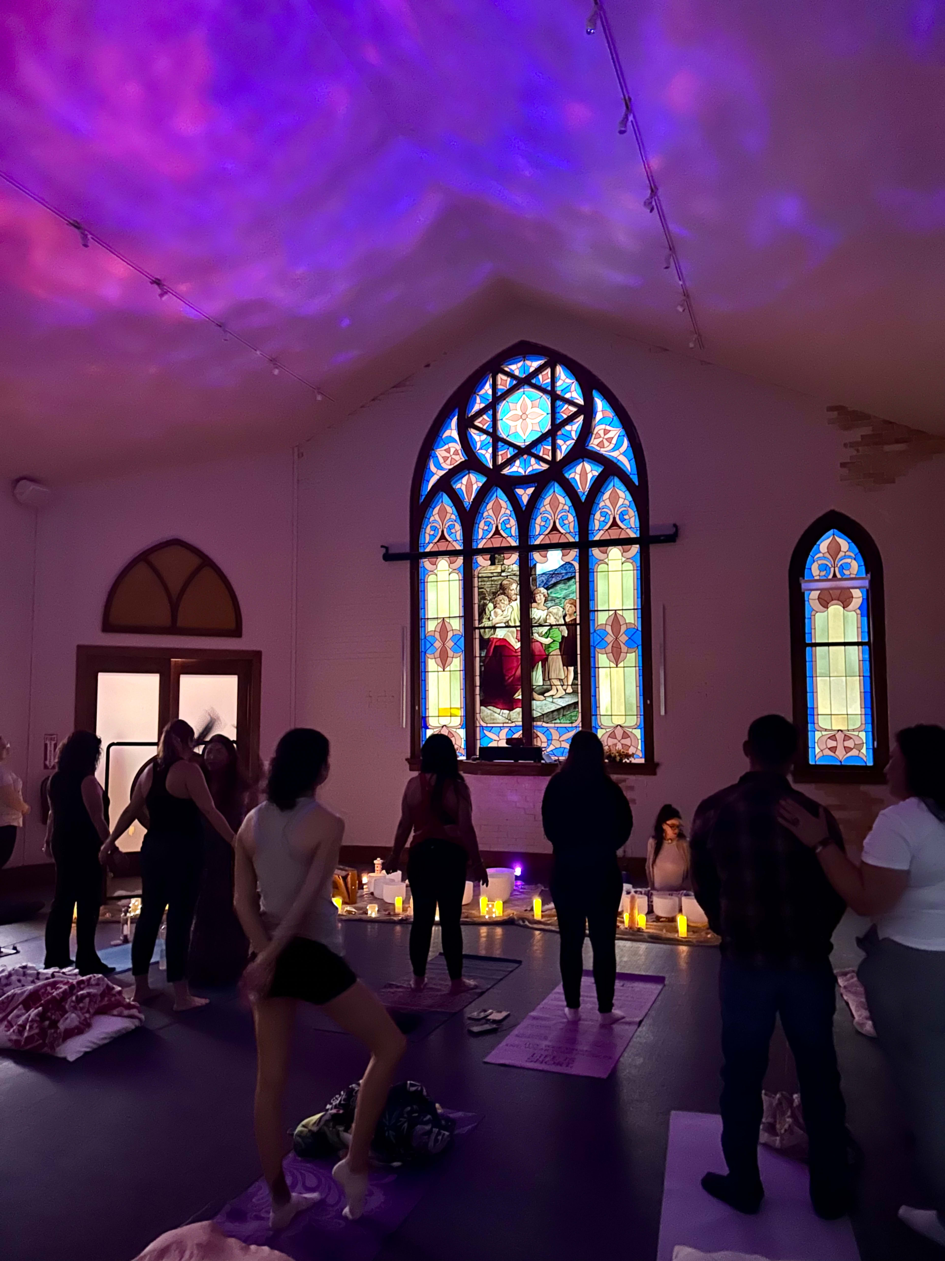 A group of people practices yoga in a dimly lit space adorned with a large stained glass window and candles.