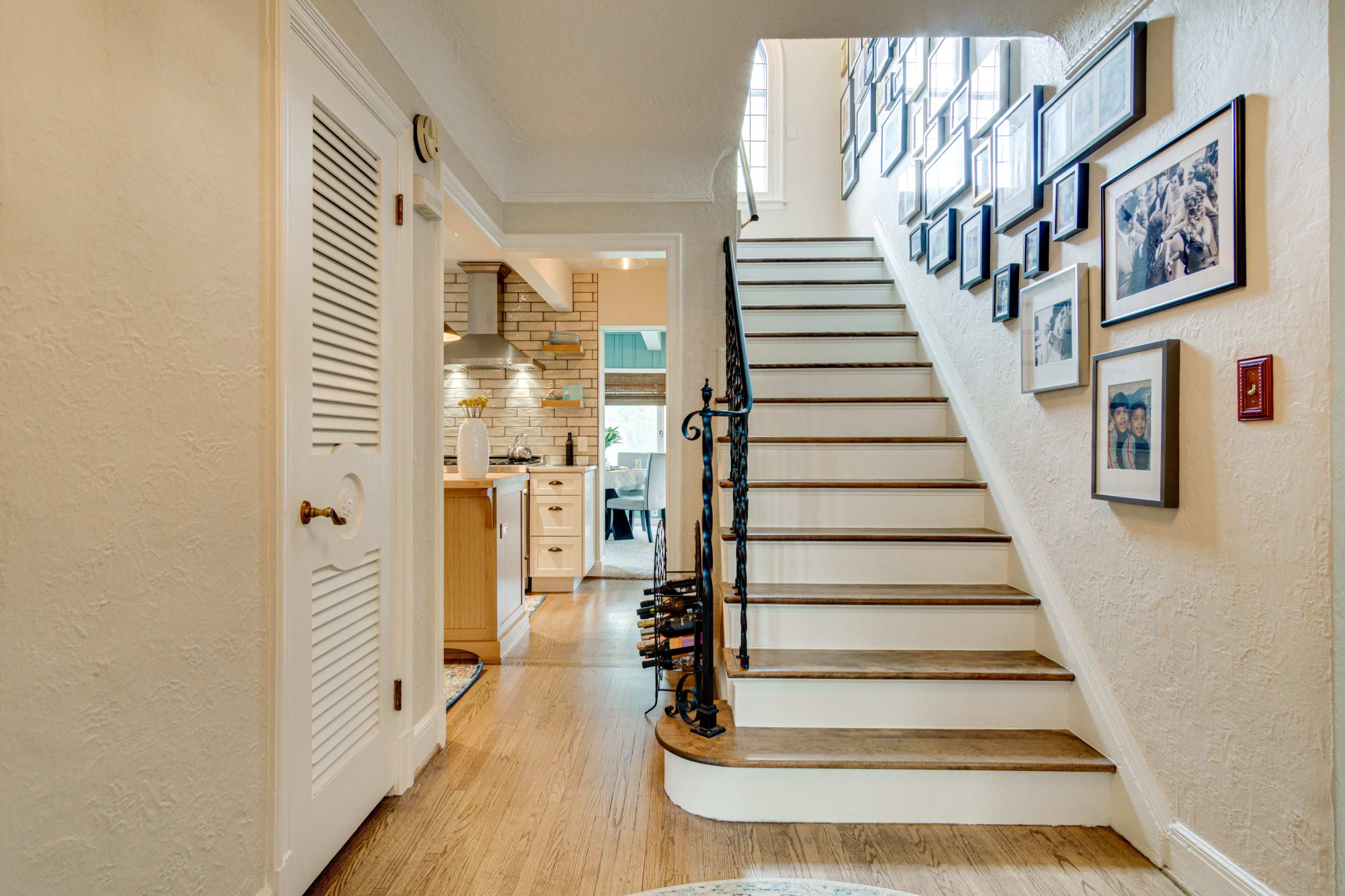 The image shows a hallway with a staircase leading to an upper level, flanked by framed photographs on the wall and a view of a kitchen in the background.
