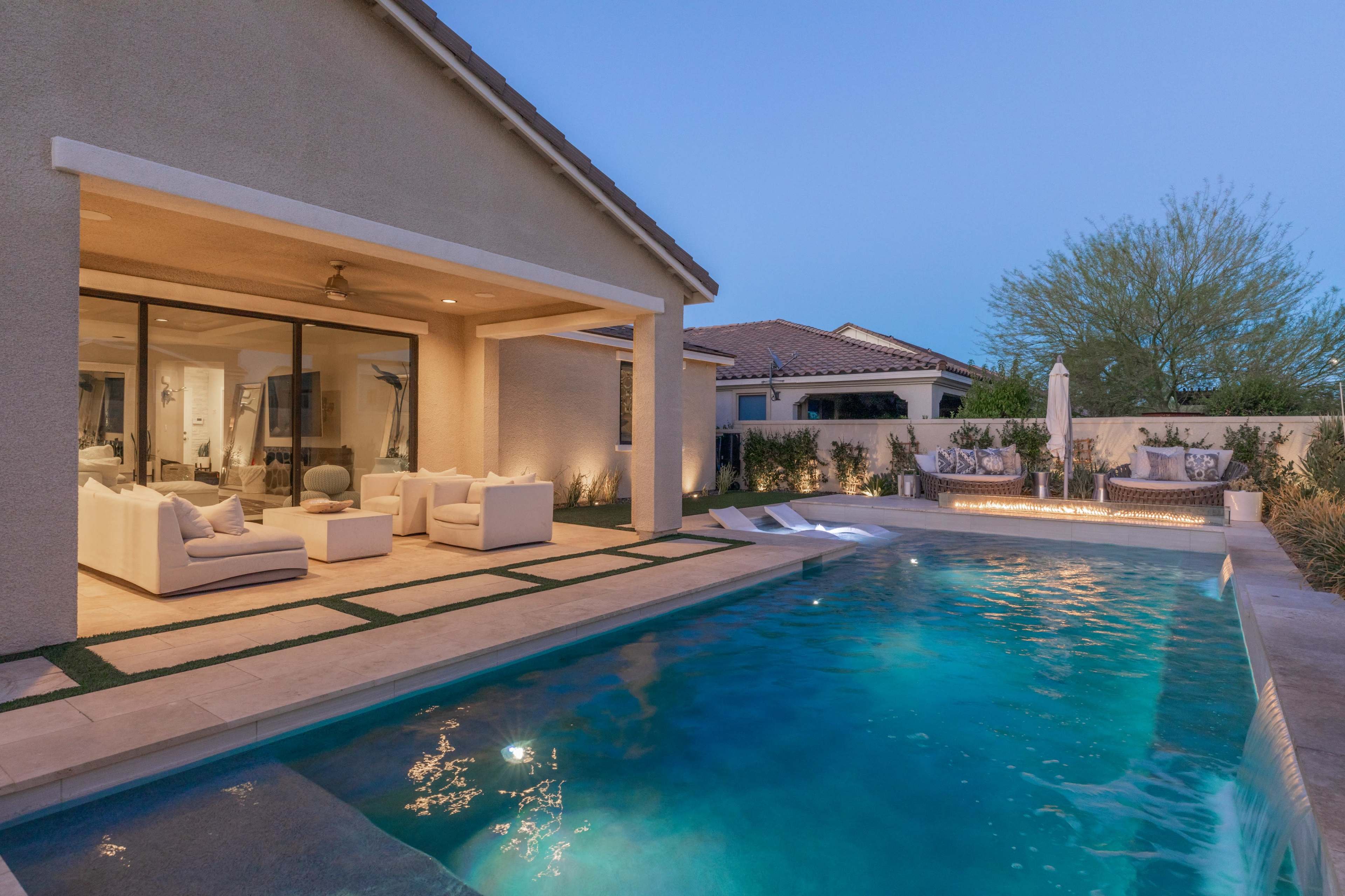 A modern house features a covered patio with seating and a swimming pool illuminated by soft lighting in the evening.