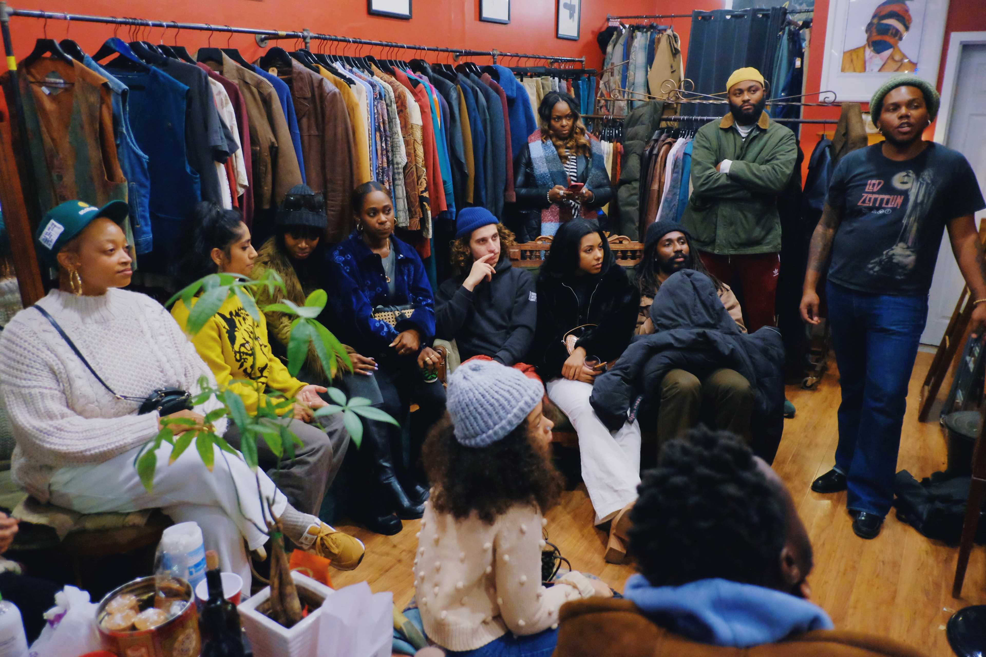 A diverse group of people is gathered in a clothing store, seated on the floor and around a small table, while one person stands and speaks to the group.