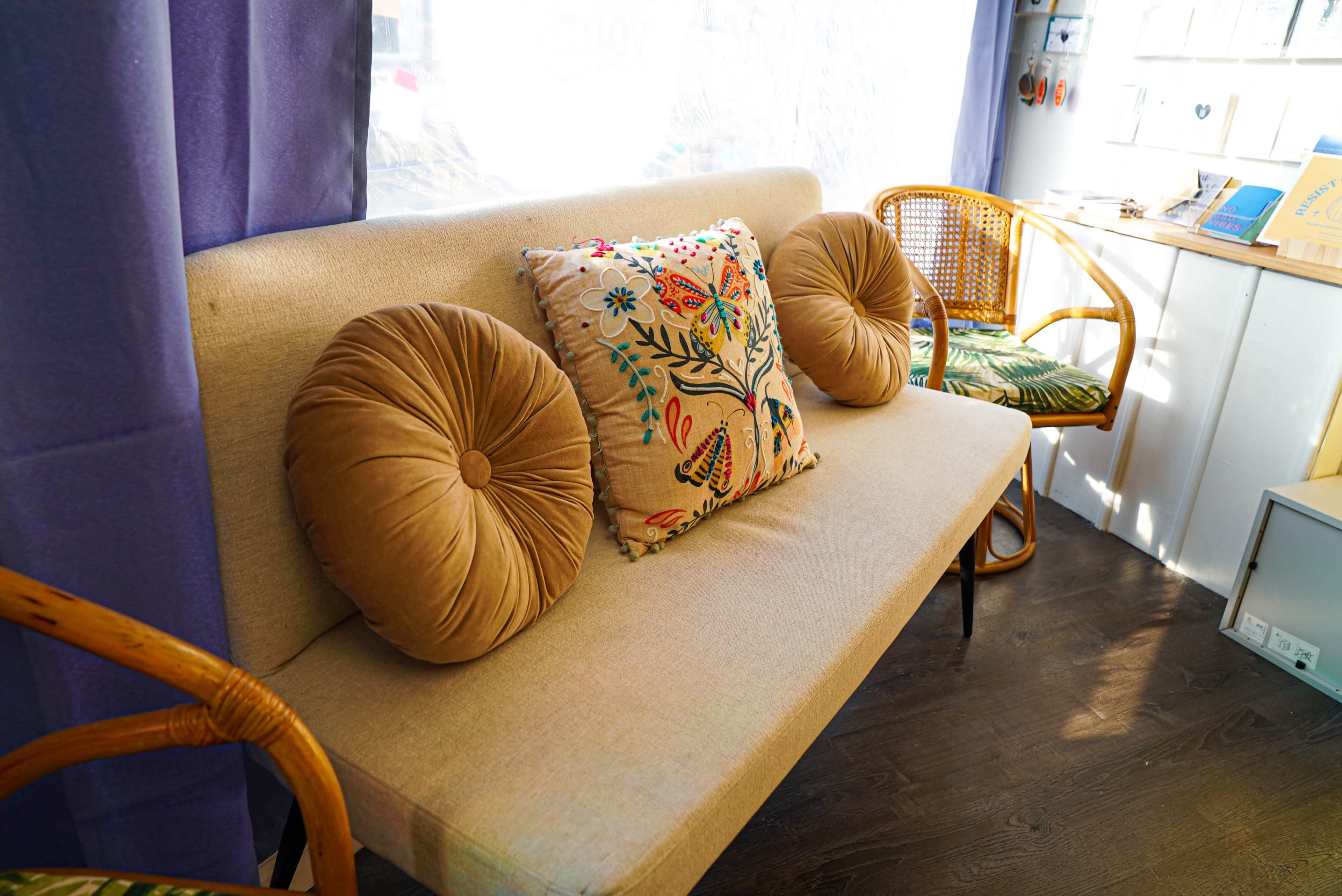The image shows a light-colored sofa with two round cushions and a decorative pillow, placed against a window with soft natural light.