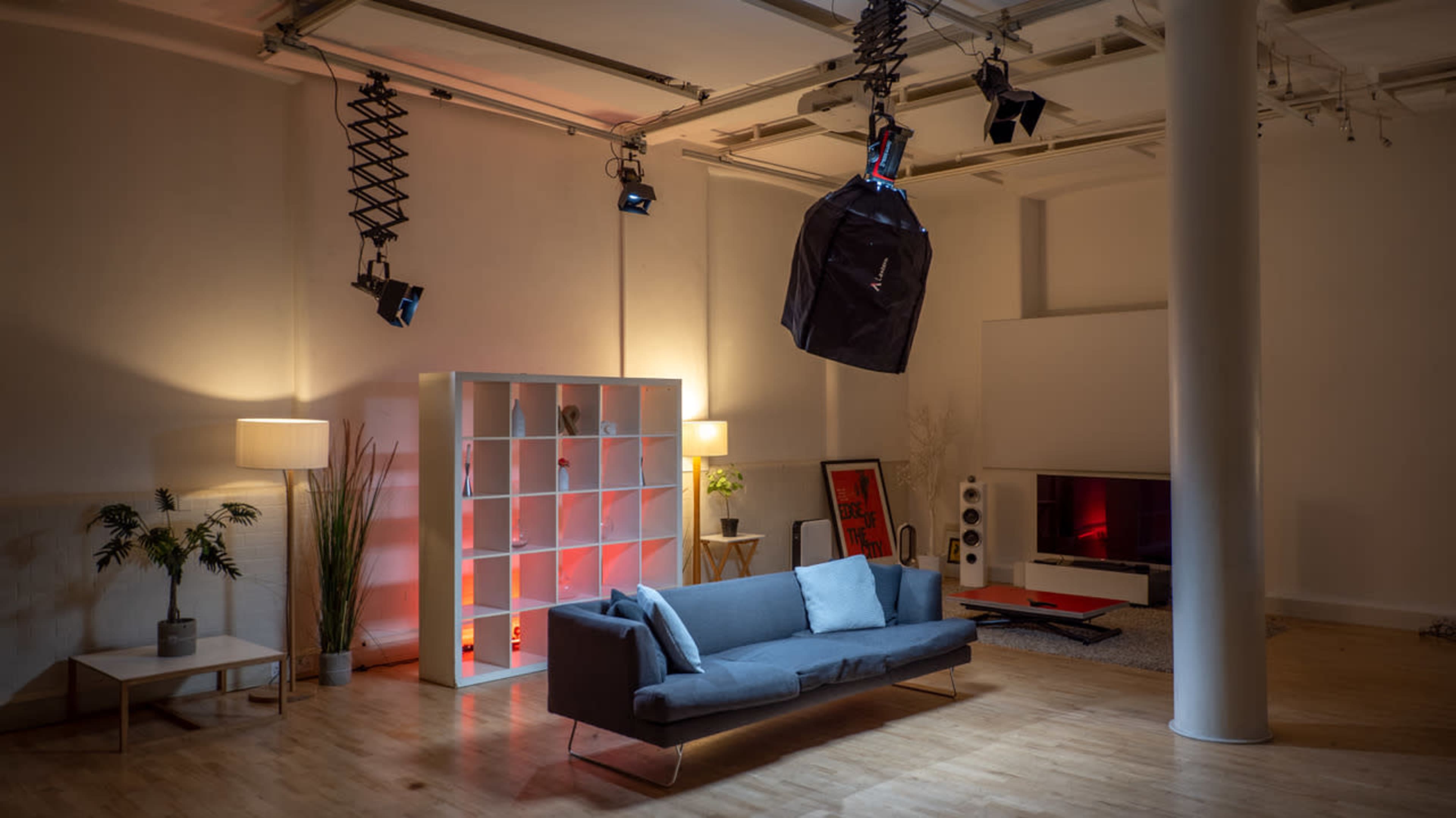 A modern living room features a gray sofa, a lighted shelving unit, a large flat-screen TV, and a suspended equipment bag in an industrial-style space.