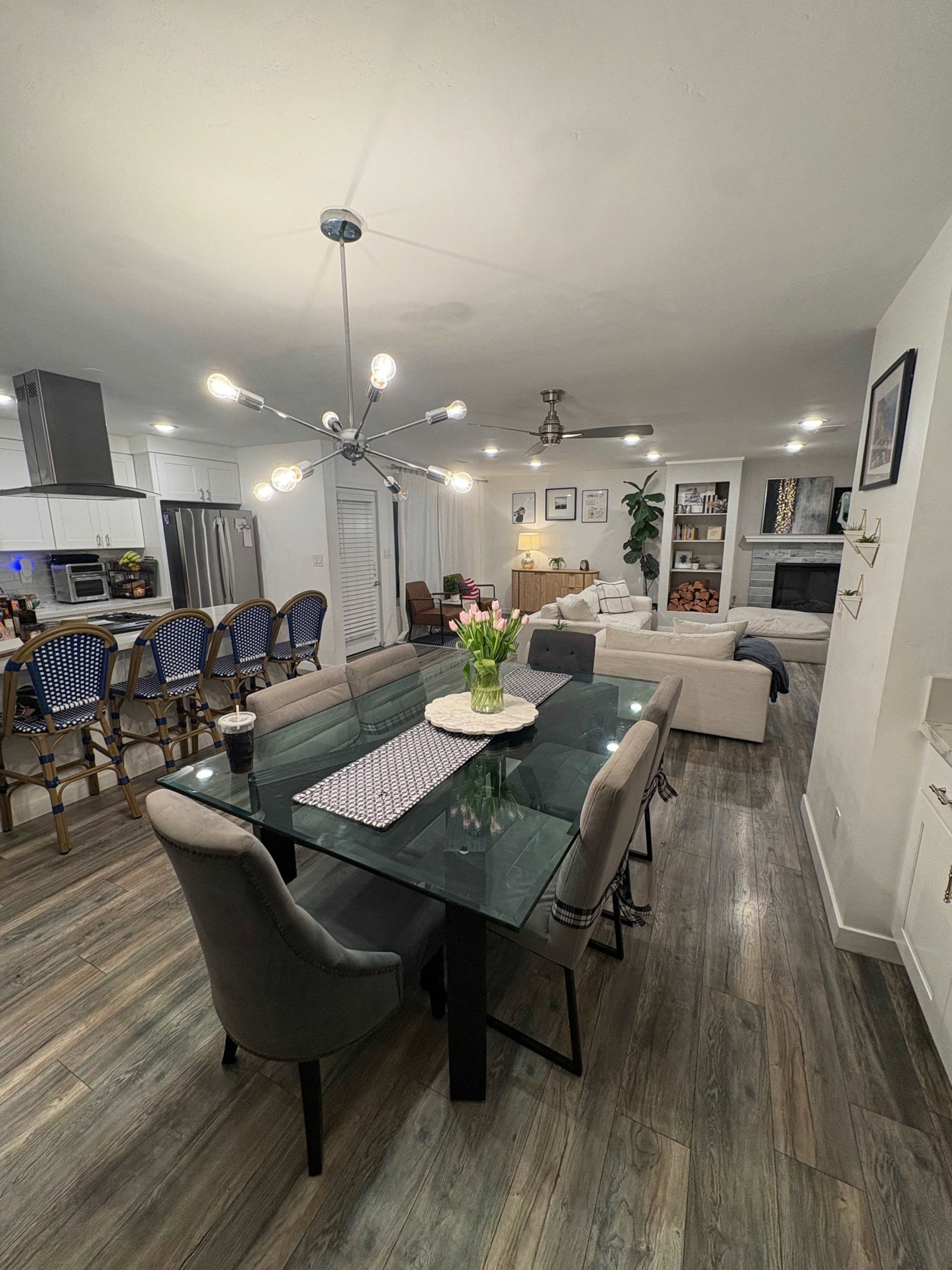 A modern dining area features a glass table surrounded by upholstered chairs, with a kitchen and living room visible in the background.