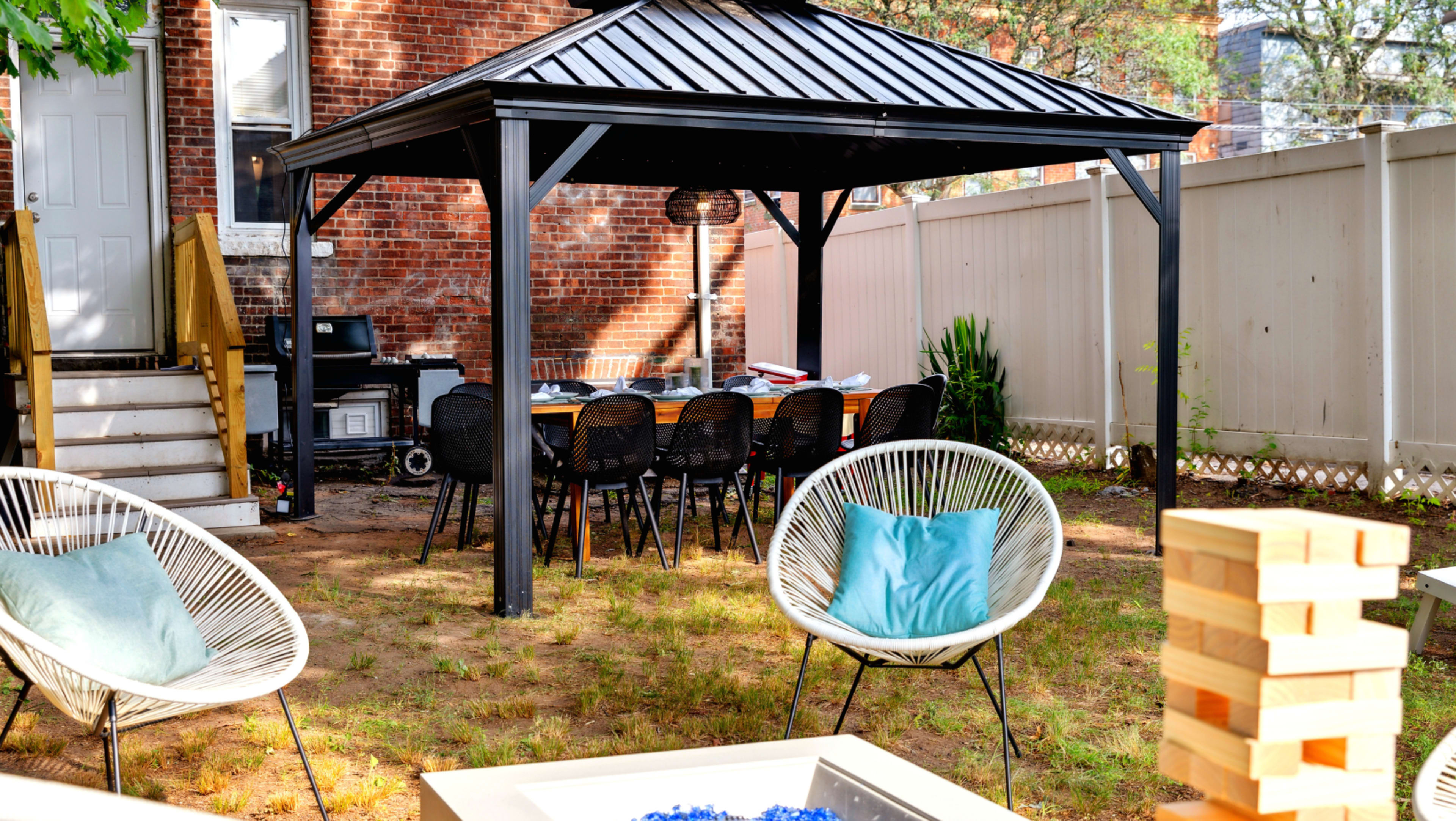 A backyard features a black gazebo with a long table and chairs underneath, surrounded by a grassy area and a seating arrangement with white chairs.