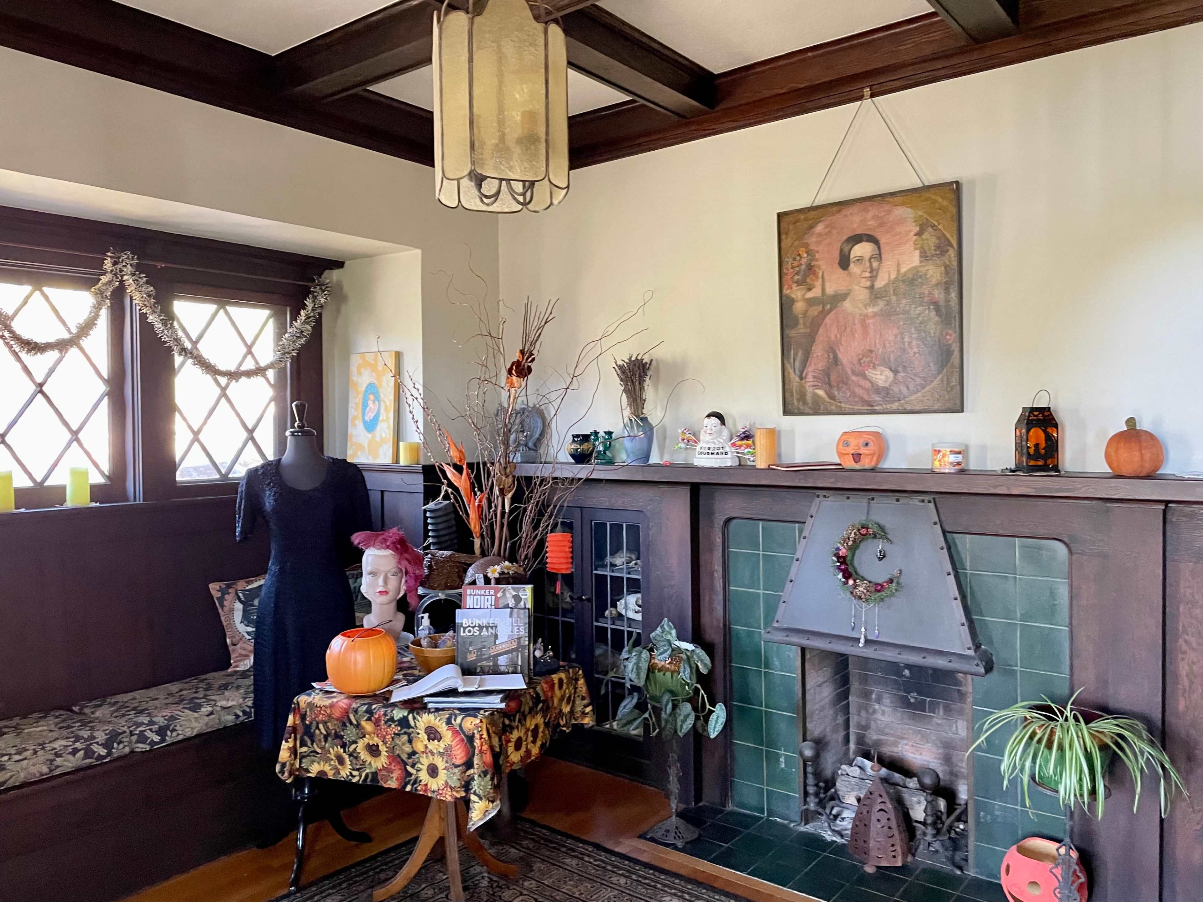 A cozy room features a fireplace with decorative pumpkins, a wooden table adorned with books and autumnal decorations, and a mannequin dressed in black, all set against a backdrop of windows with diamond-shaped panes.