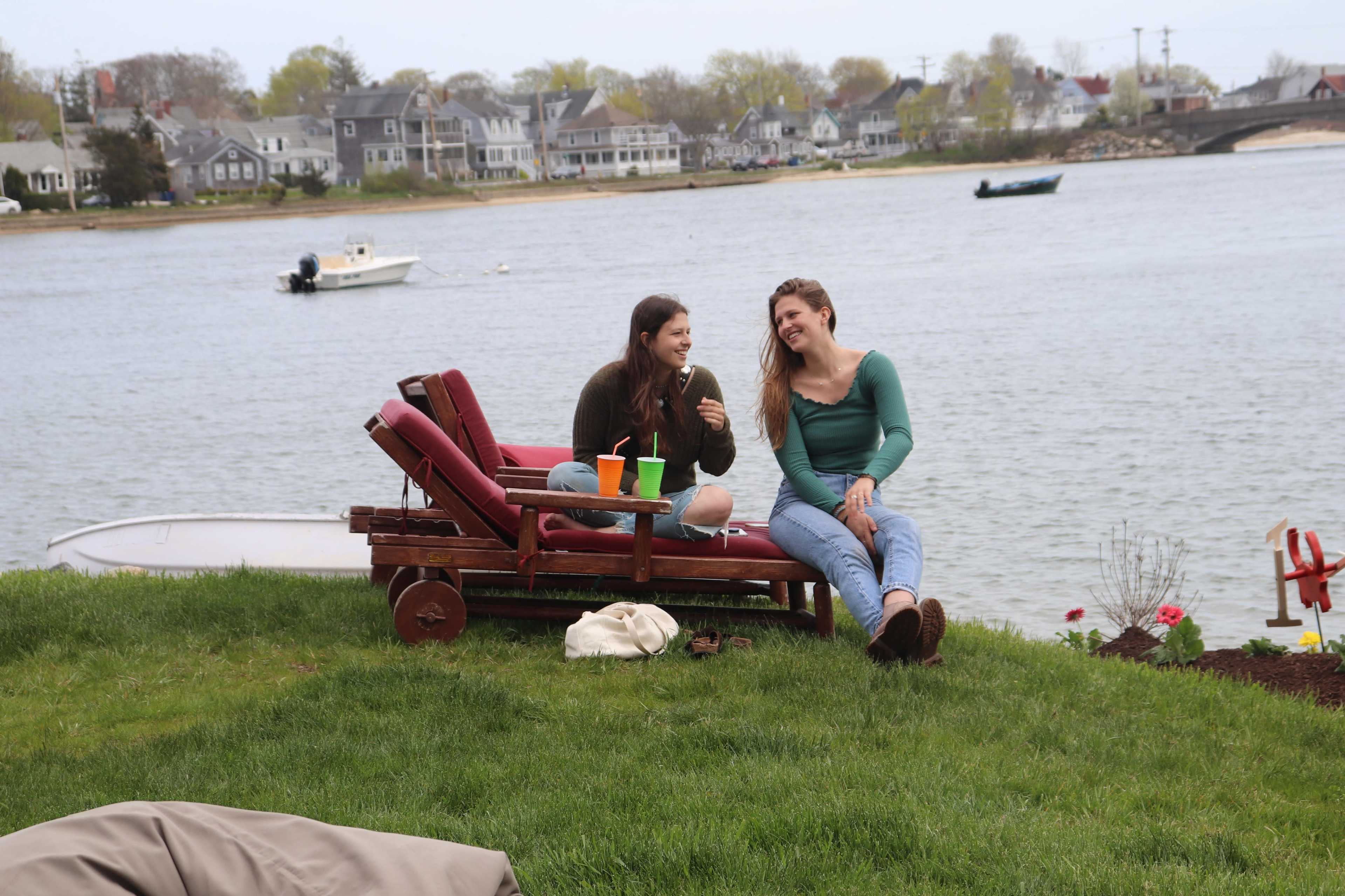 Two young women sit on a wooden lounge chair by the water, laughing together while colorful drinks are placed beside them.