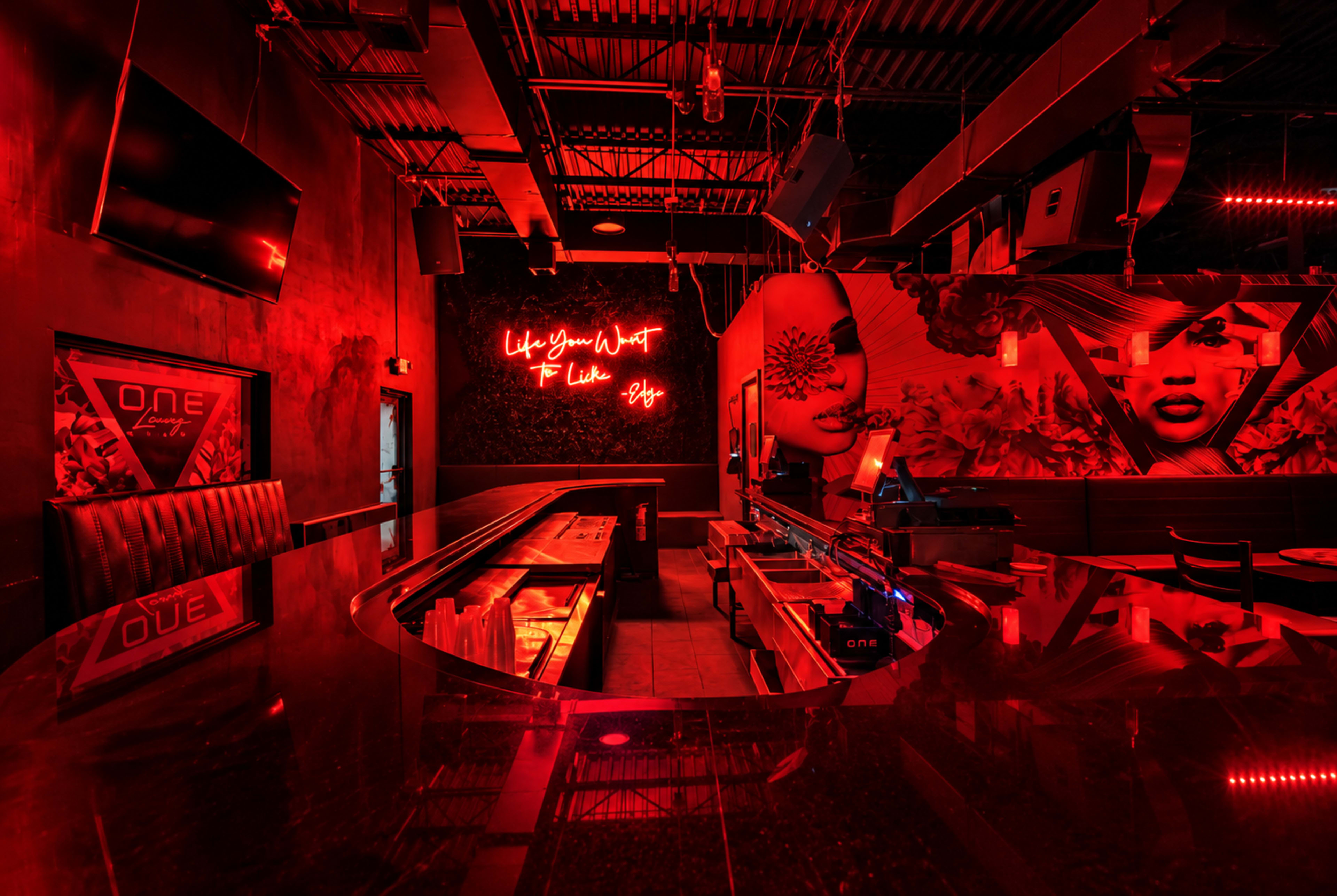 The interior of a dimly lit bar features a striking red neon sign against a vibrant mural and a sleek counter with low seating.