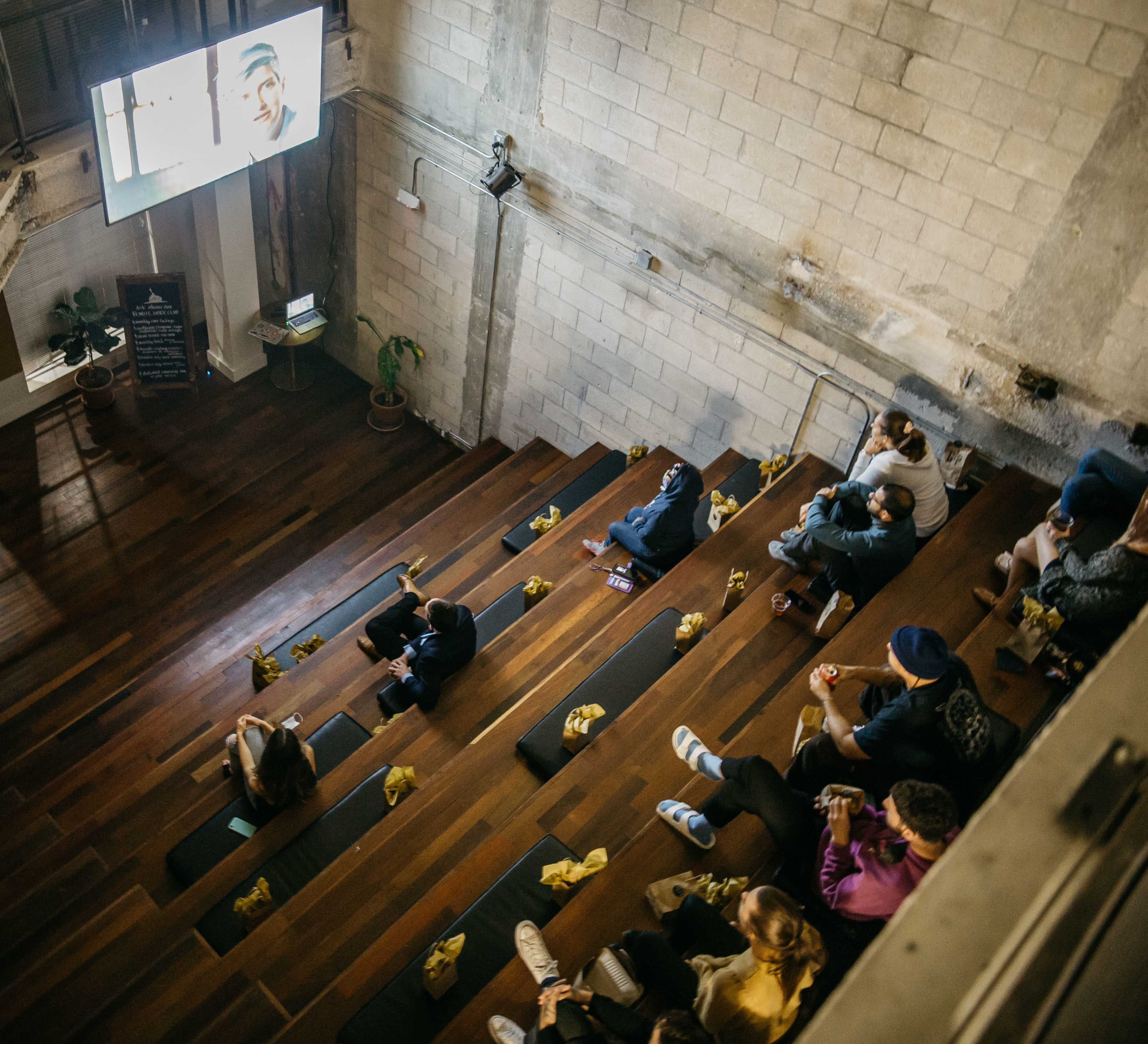 A group of people watches a presentation on a large screen in a tiered seating area made of wooden planks.